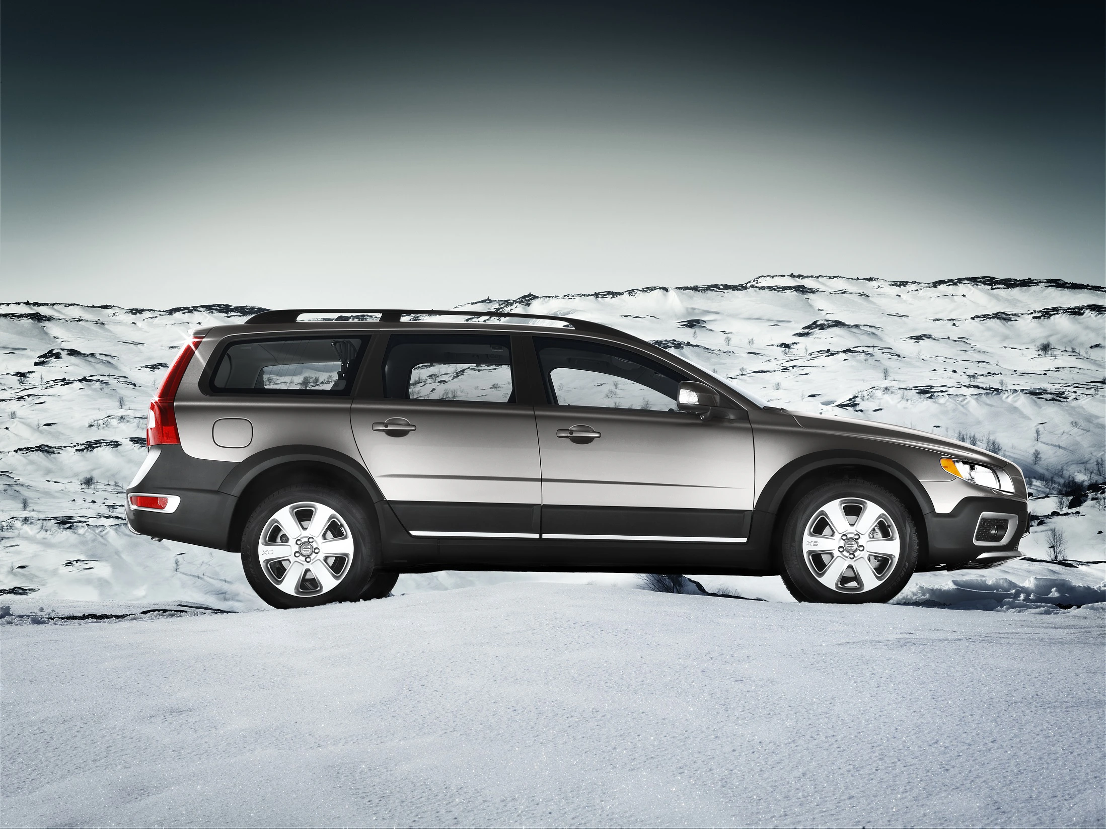 Silberner SUV parkt auf schneebedecktem Untergrund vor einem Bergpanorama unter bewölktem Himmel.