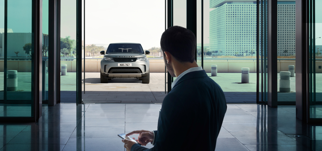 Ein Mann im Anzug steht in einem modernen Gebäude und blickt auf ein geparktes Auto im Freien. Er hält ein Tablet in der Hand.