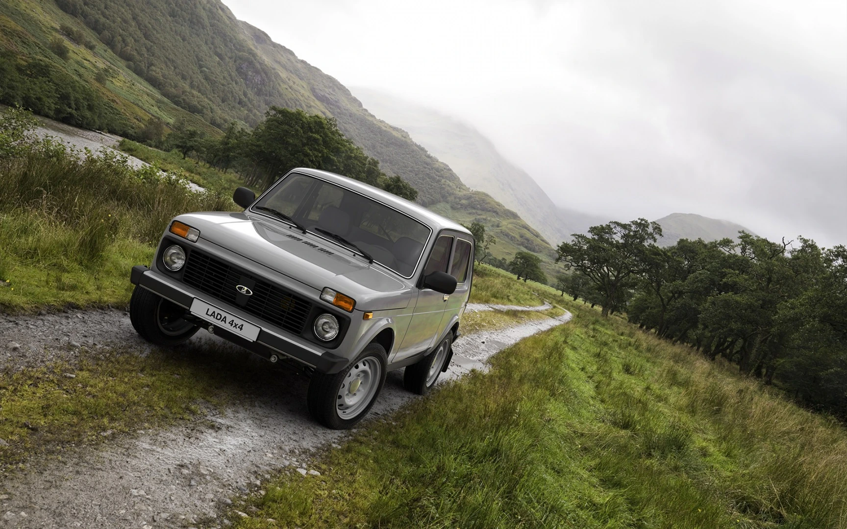 Ein silberner Lada 4x4 steht auf einem schmalen, kurvigen Pfad in einer grünen, hügeligen Landschaft unter bewölktem Himmel.