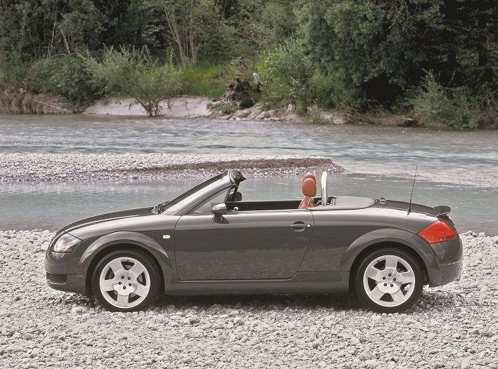 Schwarzes Cabriolet auf Kiesstrand, neben einem Fluss, umgeben von grünen Bäumen. Autodach ist geöffnet, zeigt roten Innenraum.