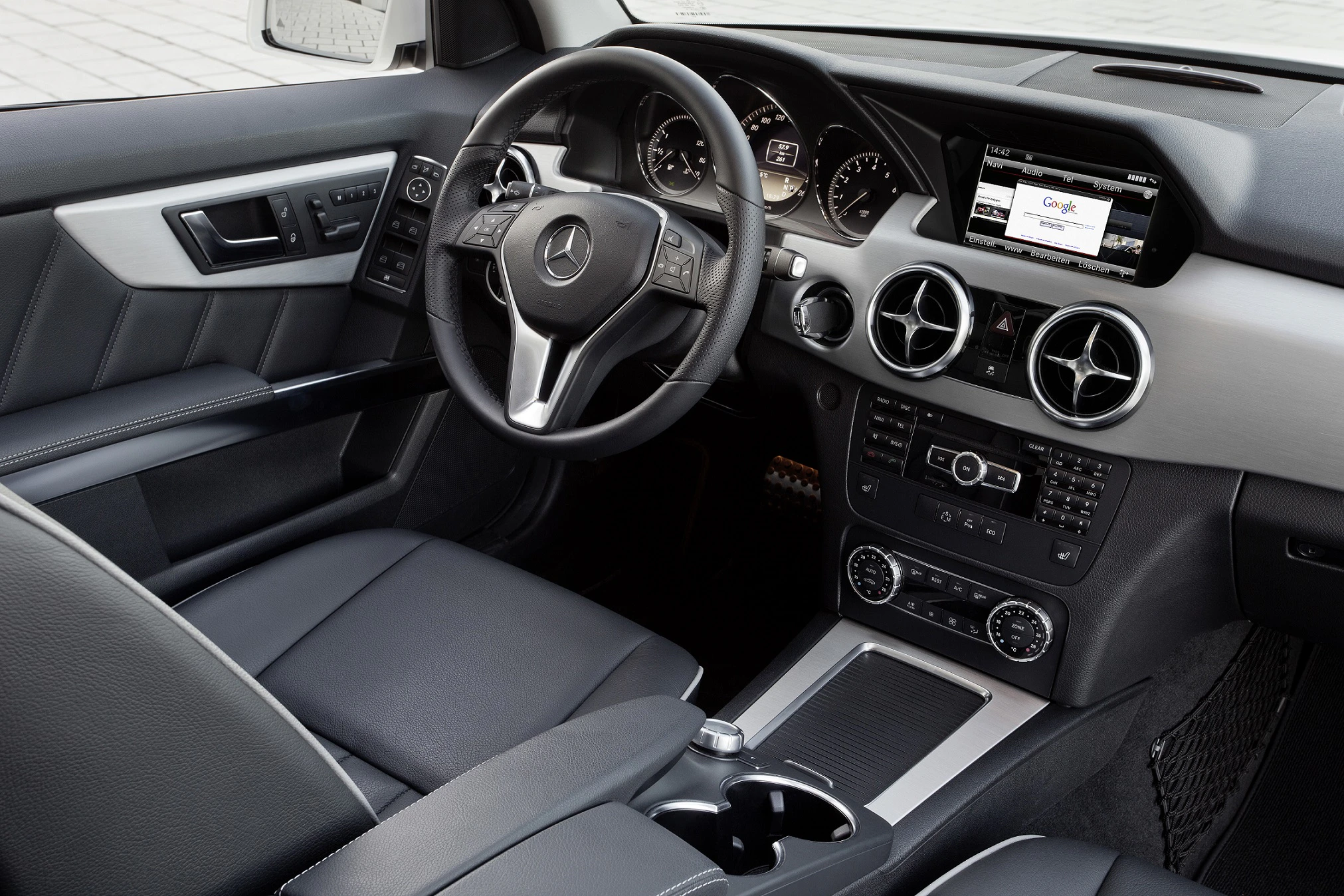Innenraum eines Mercedes mit Lederpolstern, Lenkrad mit Logo, Armaturenbrett mit Bildschirm und Anzeigen, Mittelkonsole mit Bedienelementen.