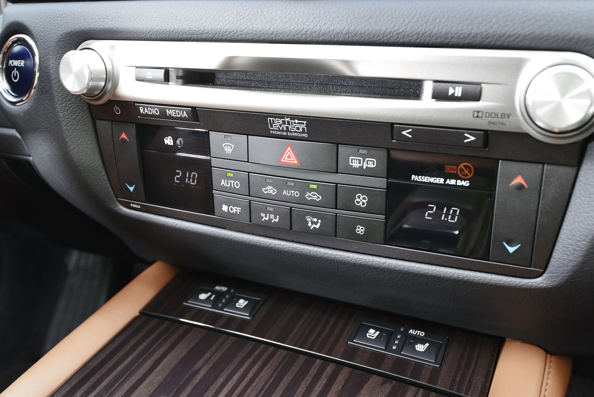 Steuerkonsole eines Autos mit Klimaanlage, Radio, Medien und Bedienelementen. Holzverkleidung und Ledersitz erkennbar.
