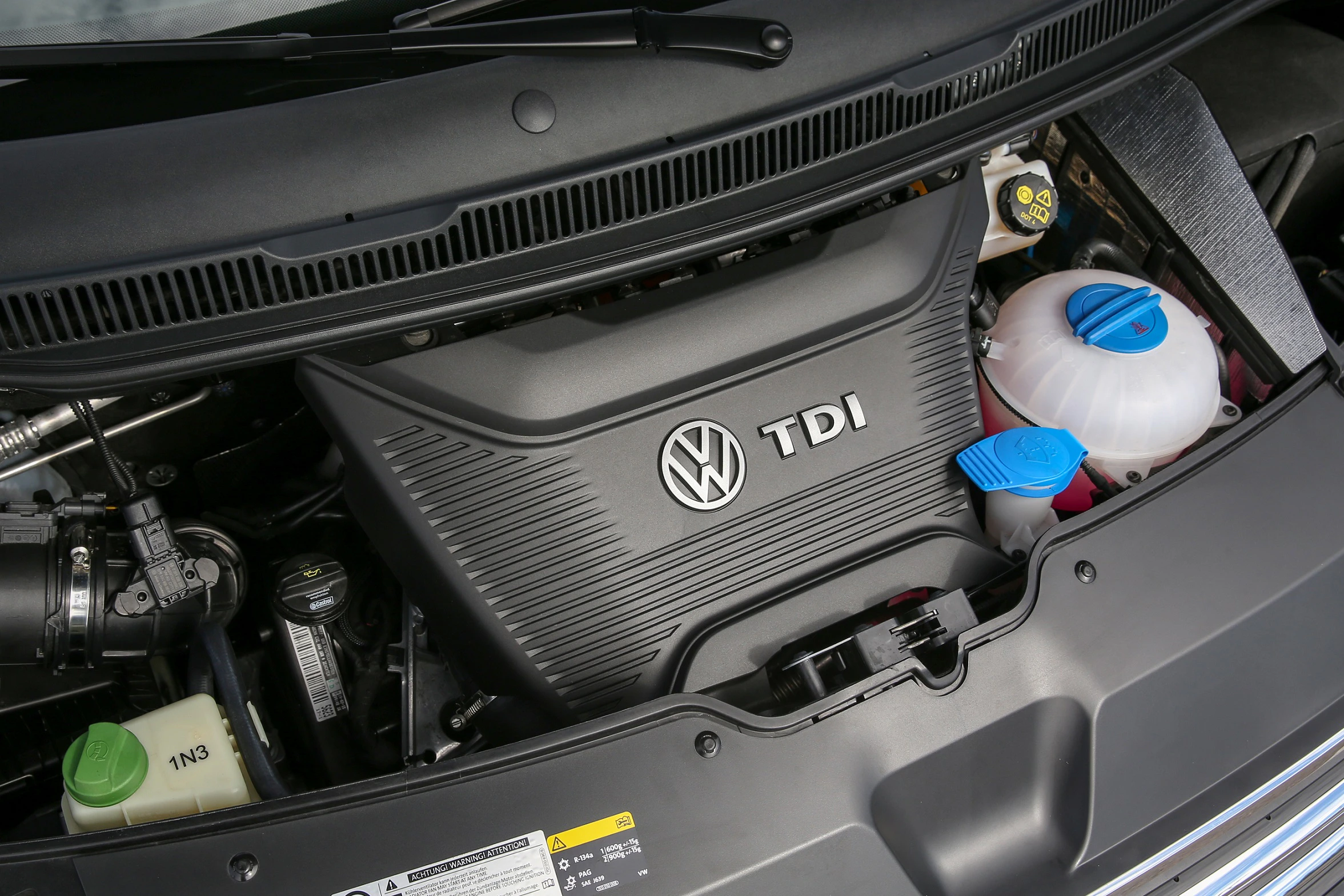 Nahaufnahme eines VW TDI-Motors unter einer geöffneten Motorhaube, mit sichtbaren Flüssigkeitsbehältern und Motorkomponenten.