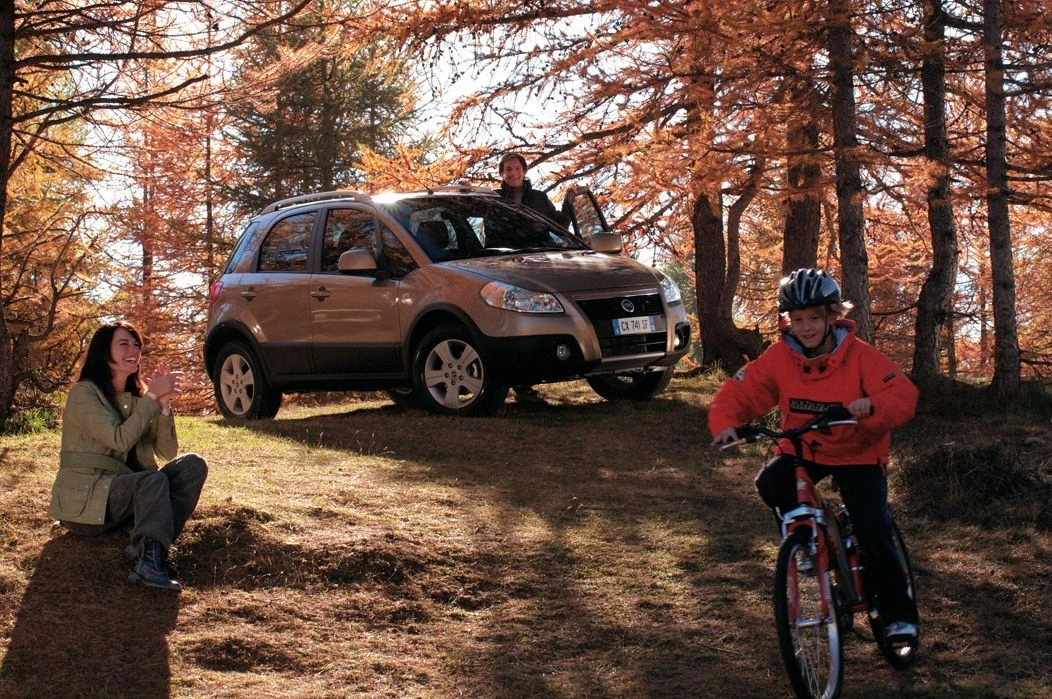 Eine Familie genießt einen herbstlichen Tag im Wald; ein Kind fährt Fahrrad, während Eltern in der Nähe eines geparkten Autos stehen und sitzen.