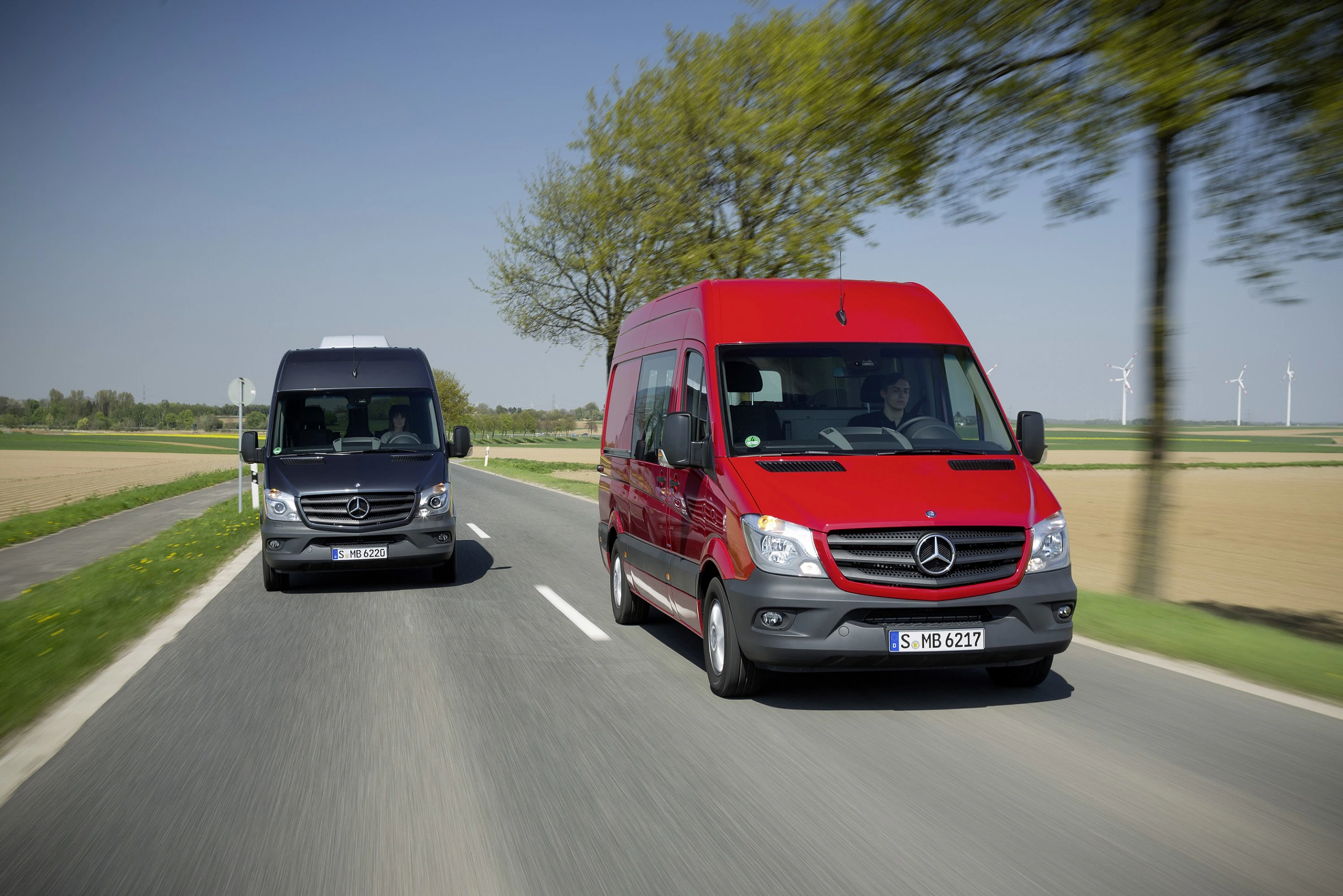 Zwei Mercedes-Transporter fahren auf einer Landstraße; der rote vorne und der schwarze hinten. Windräder und Felder im Hintergrund.