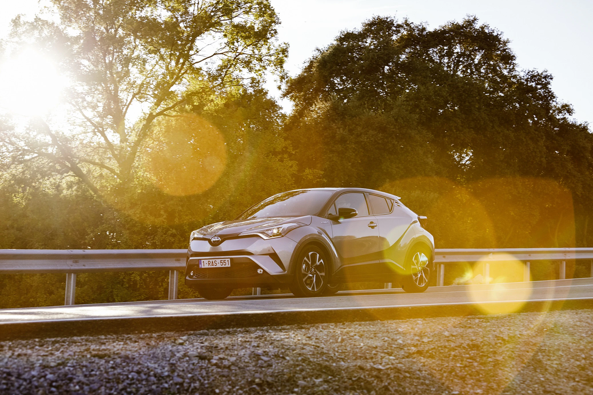 Silberner SUV fährt auf Straße bei Sonnenuntergang, umgeben von Bäumen. Sonnenstrahlen und Lens Flare-Effekt im Bild.