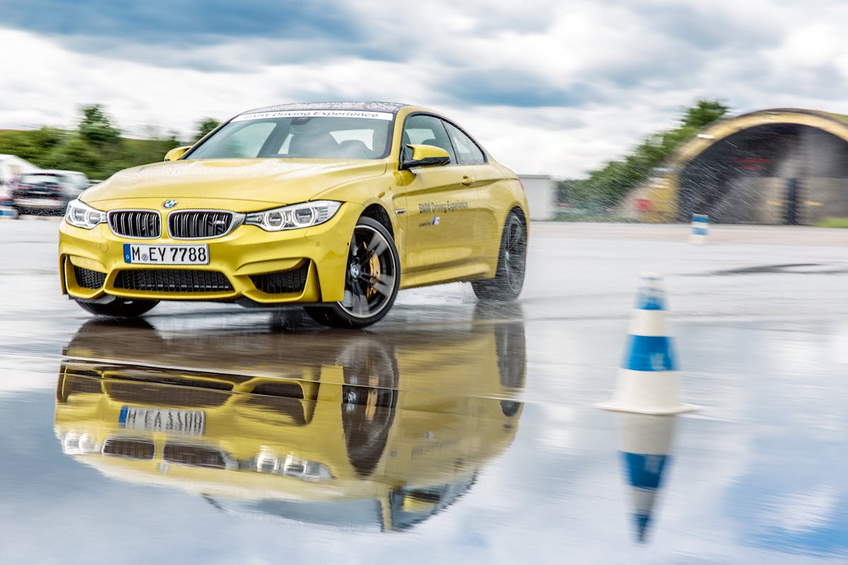 Gelber BMW auf nasser Fahrbahn, driftet zwischen blauen Kegeln. Spiegelung des Autos im Wasser. Hintergrund mit Wolken und verstärktem Himmel.