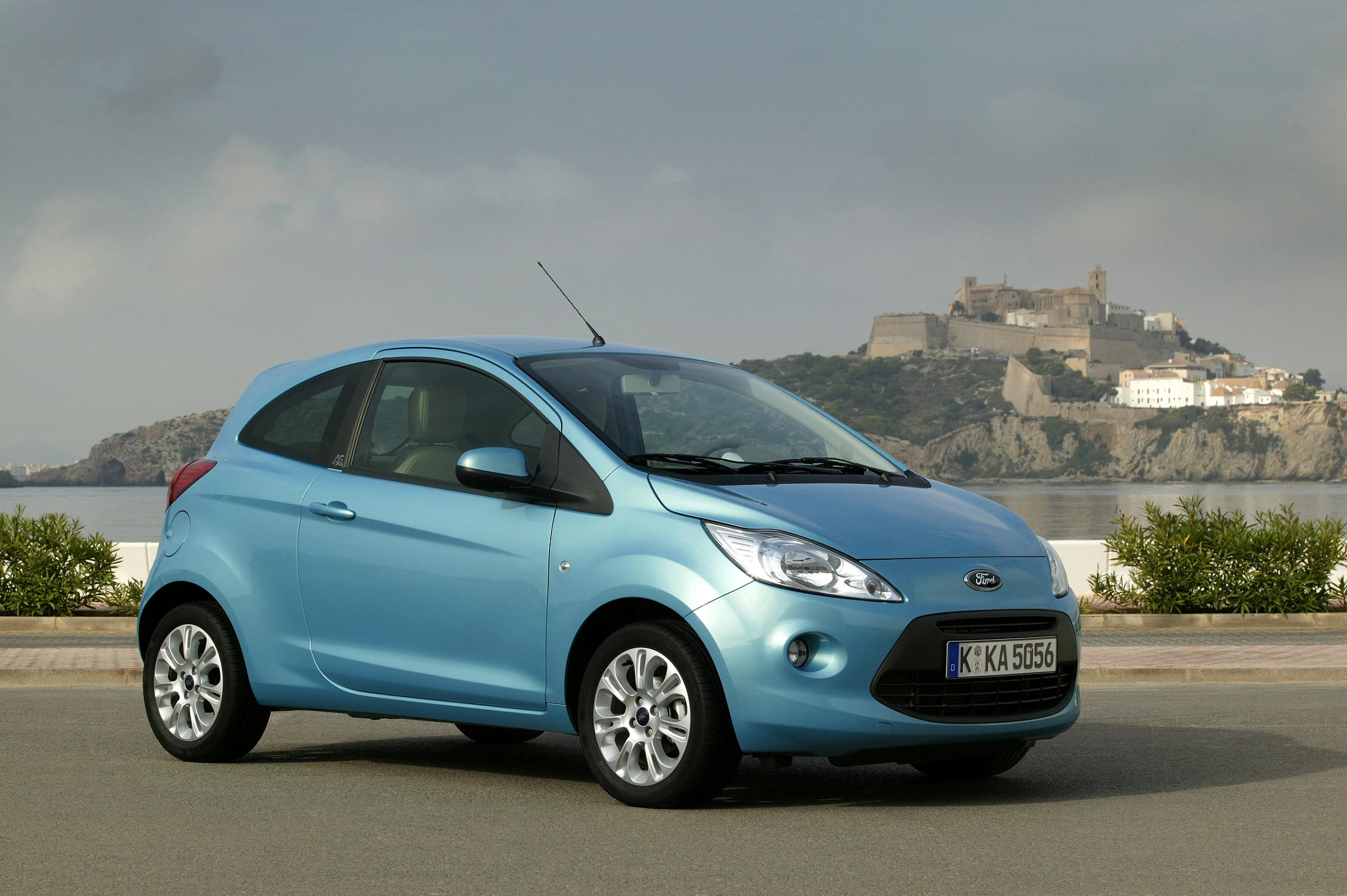 Blauer Ford-Kleinwagen parkt auf Straße vor malerischer Hügelkulisse mit Burg und bewölktem Himmel im Hintergrund.