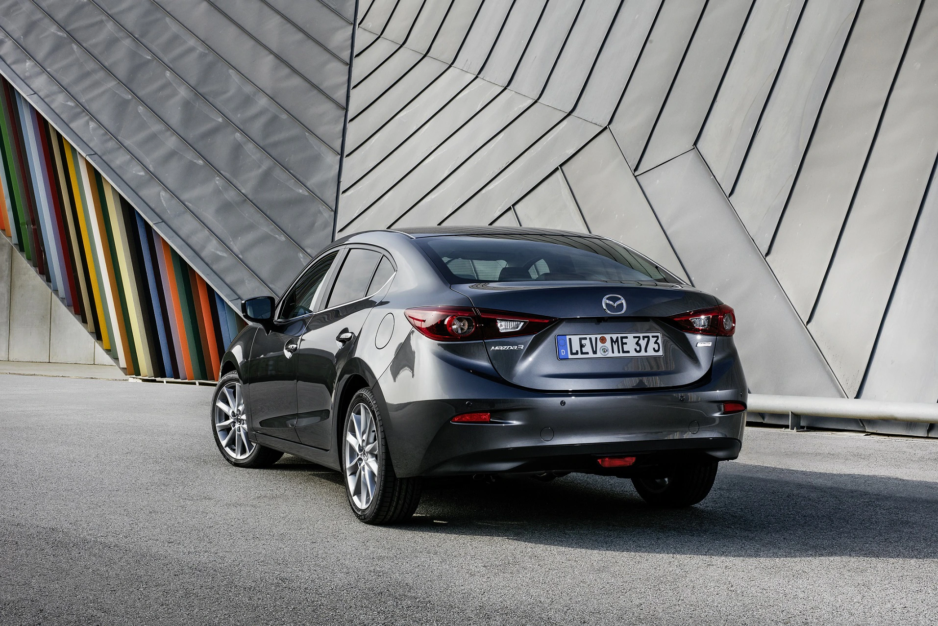 Ein grauer Mazda parkt vor einer modernen, bunten Metallfassade auf grauem Asphalt.
