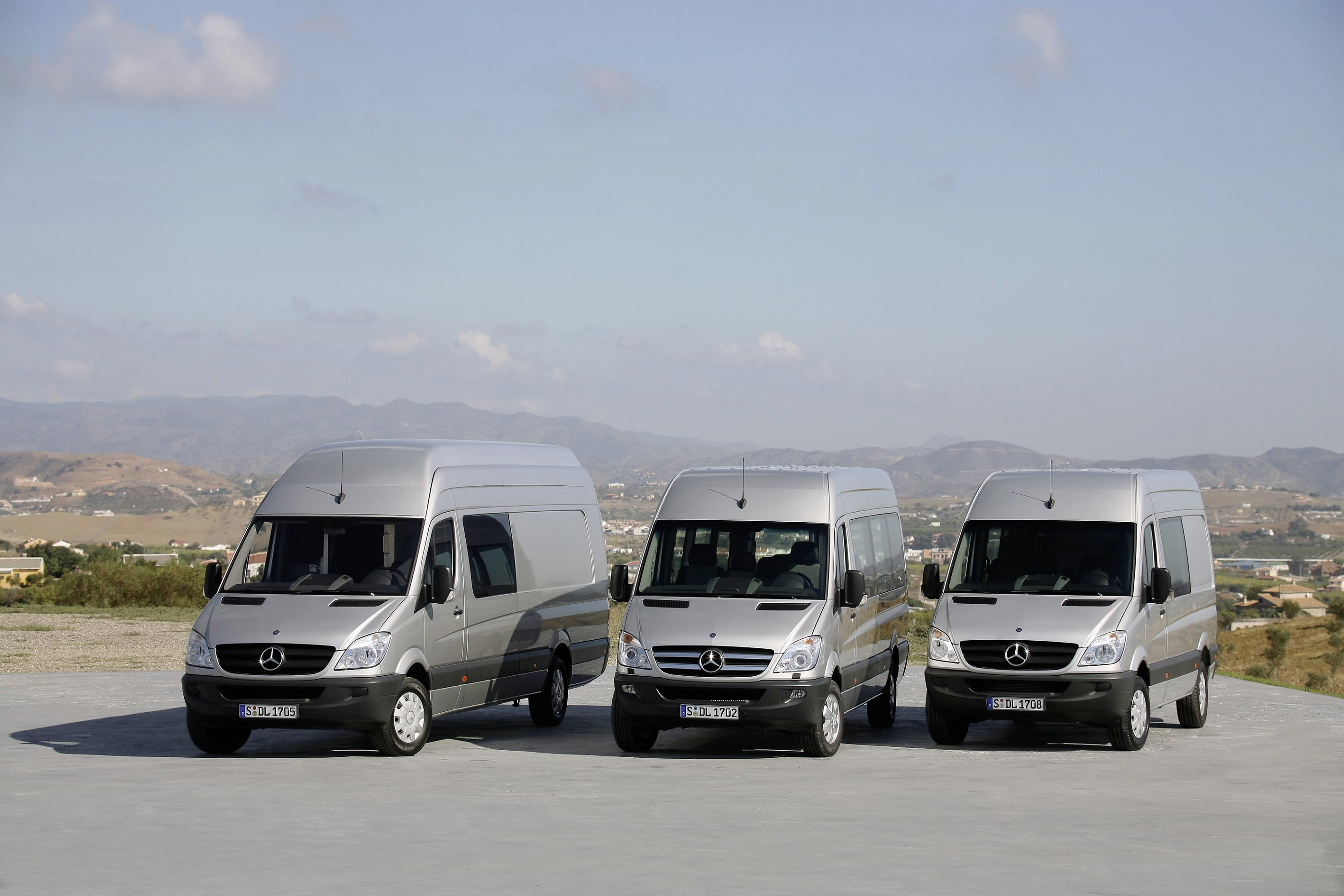 Drei silberne Mercedes-Benz Transporter stehen nebeneinander auf einer Straße mit hügeliger Landschaft im Hintergrund.