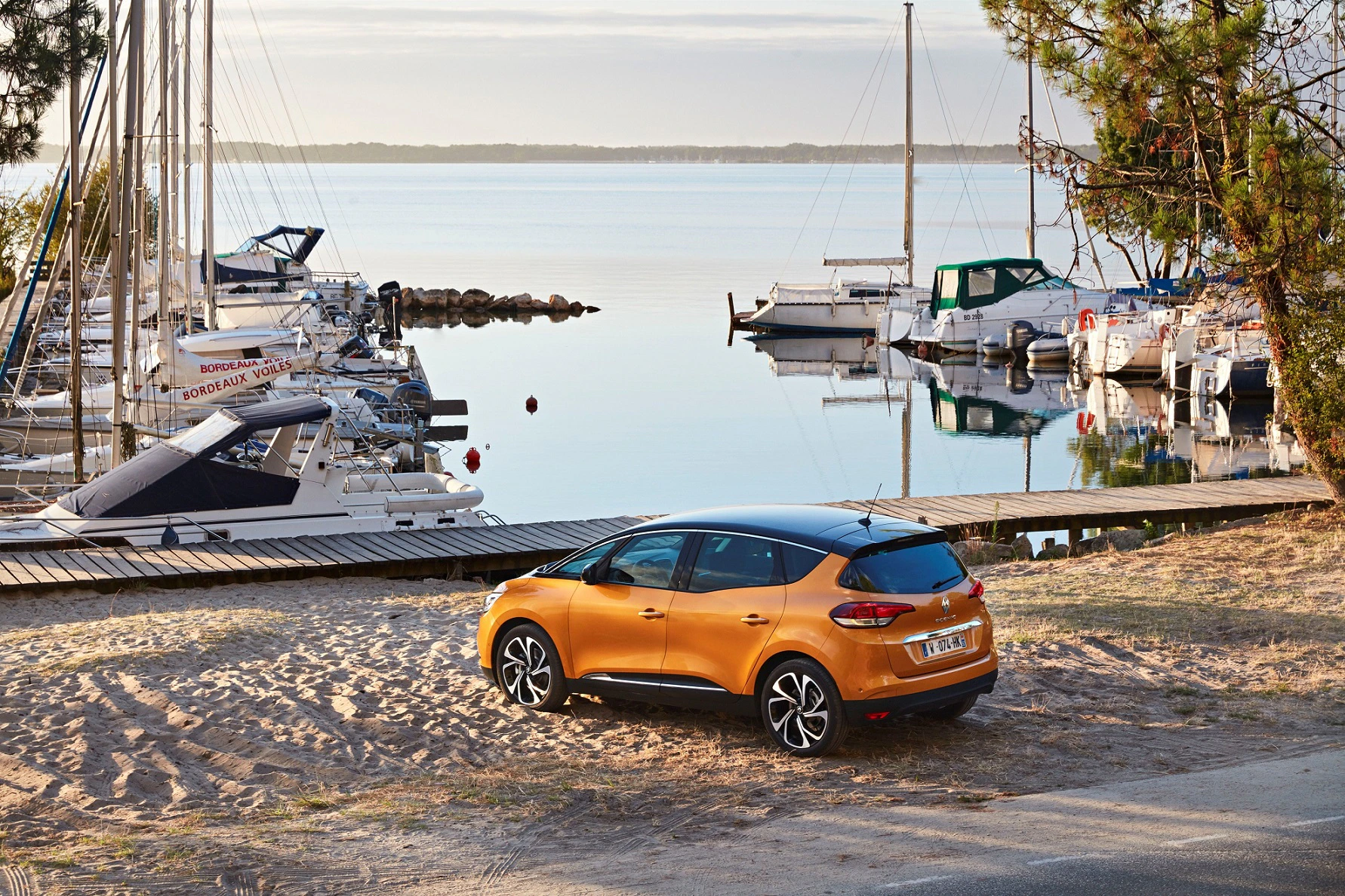 Ein orangefarbenes Auto parkt auf sandigem Boden neben einem Yachthafen mit mehreren Segelbooten, umgeben von ruhigem Wasser und Kiefern.
