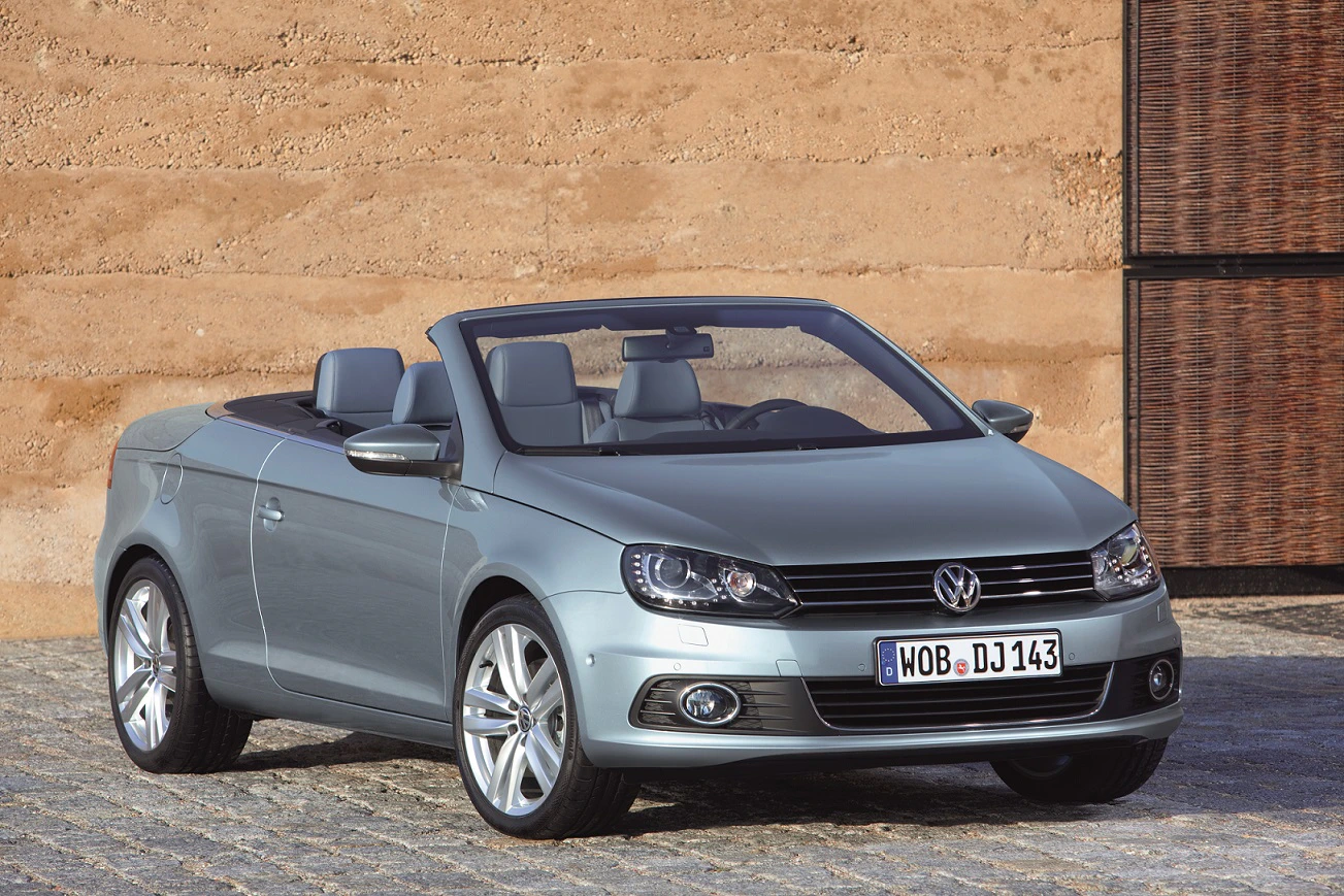 Silbernes Volkswagen-Cabriolet steht vor einer strukturierten Wand auf Kopfsteinpflaster, Verdeck geöffnet, mit glänzenden Felgen.