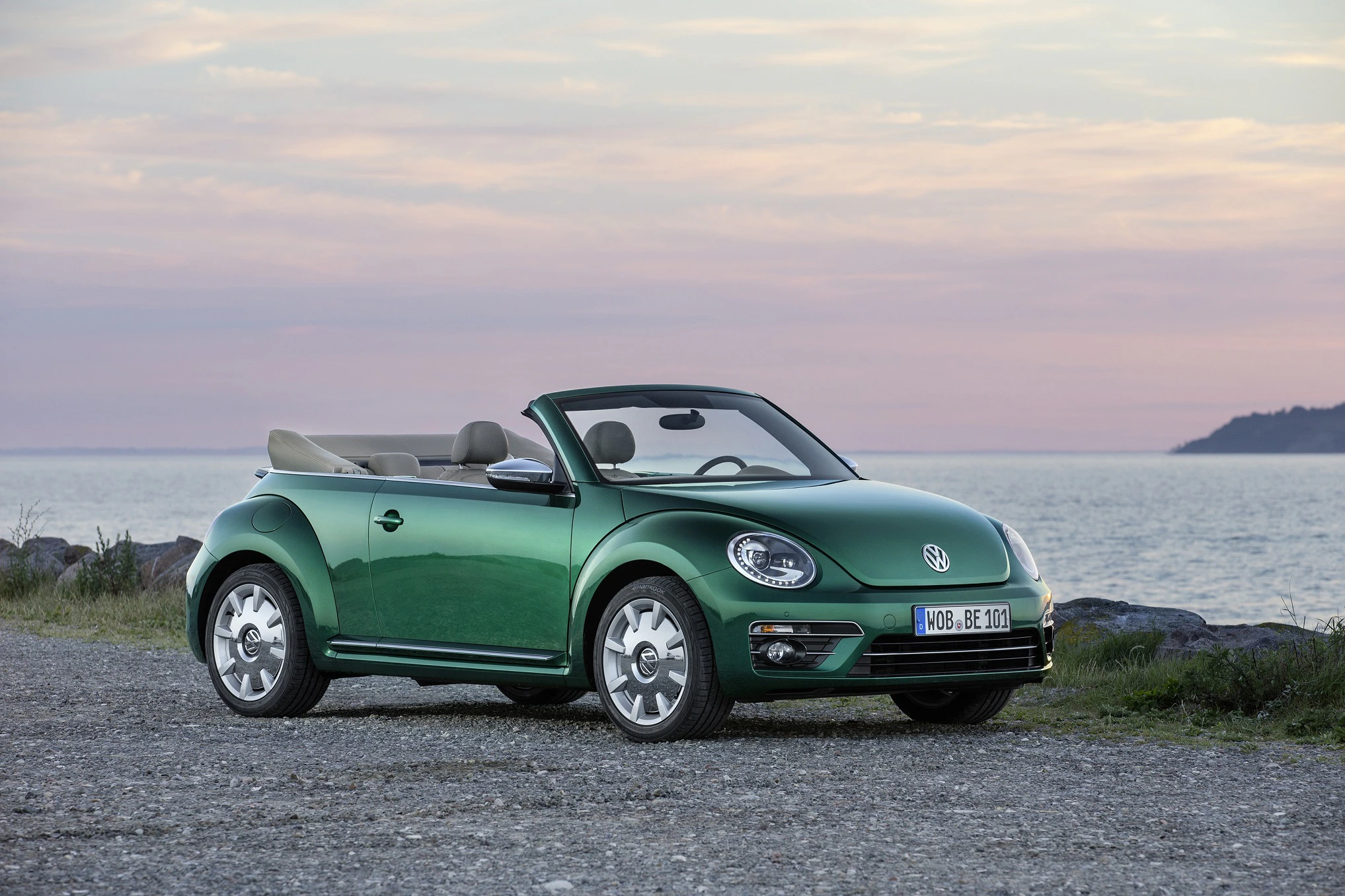 Grünes VW Beetle Cabrio steht auf Schotterweg vor einem Meer mit rosa Abendhimmel im Hintergrund. Dach ist offen, Fahrzeug von vorne seitlich sichtbar.