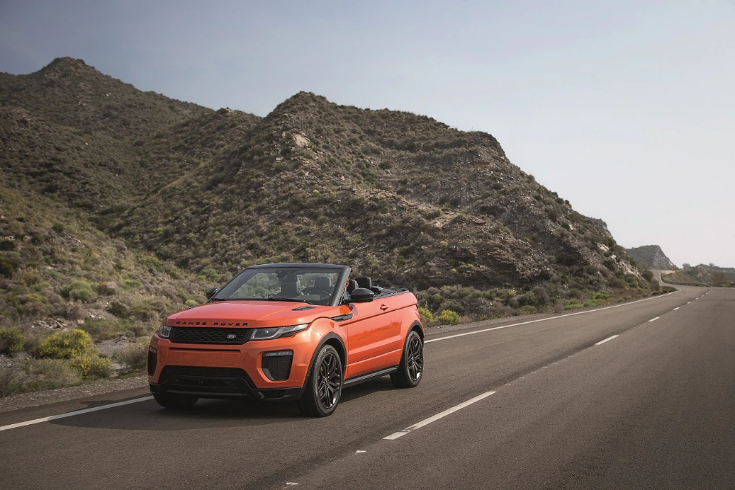 Orangefarbener Cabrio-Geländewagen fährt auf einer leeren, asphaltierten Straße durch eine bergige Landschaft unter klarem Himmel.