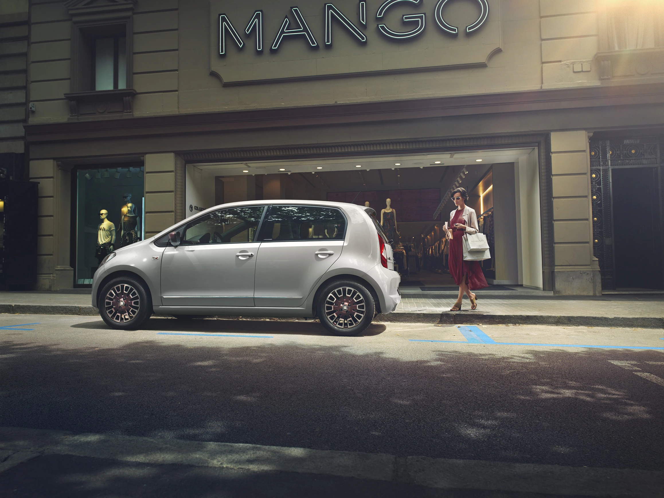 Ein silbernes Auto parkt vor einem Geschäft mit der Aufschrift „MANGO“. Eine Frau in einem roten Kleid geht am Gehweg vorbei.