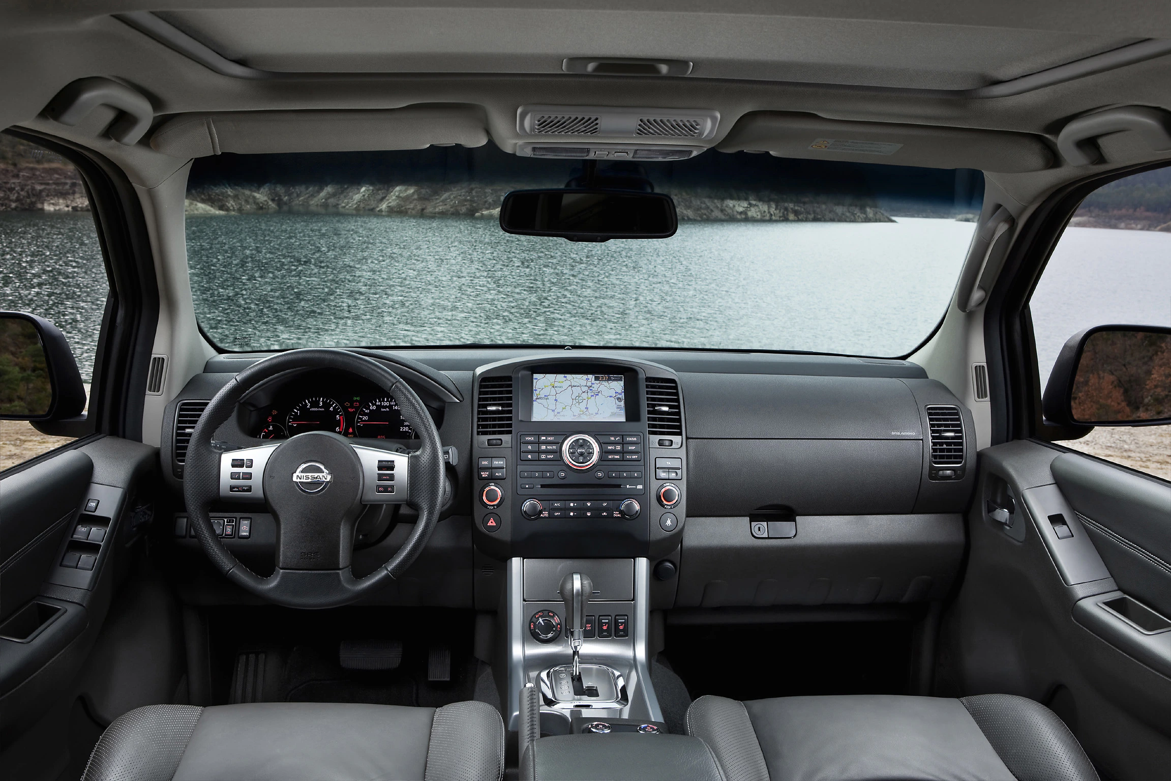 Innenraum eines Autos mit modernem Armaturenbrett, Navigationssystem und Blick durch die Windschutzscheibe auf einen See und Hügel.