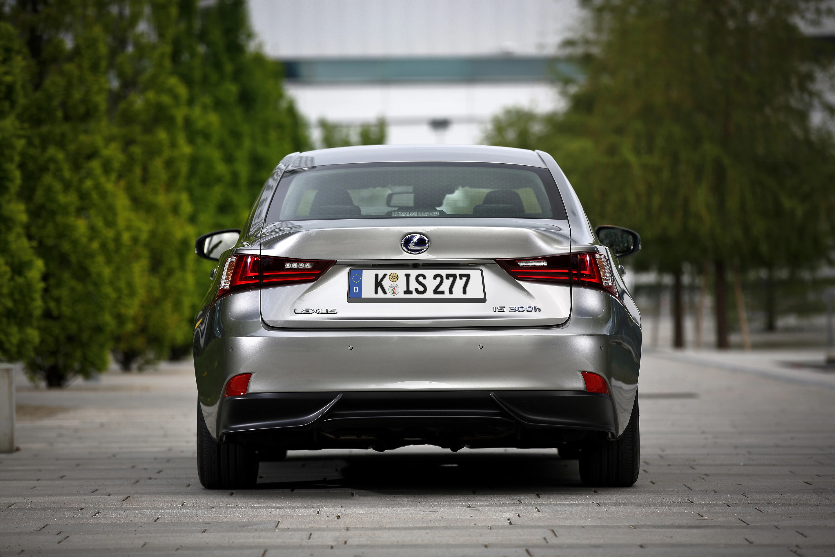 Ein silberner Lexus IS 300h, von hinten gesehen, steht auf einer gepflasterten Straße, flankiert von Bäumen.