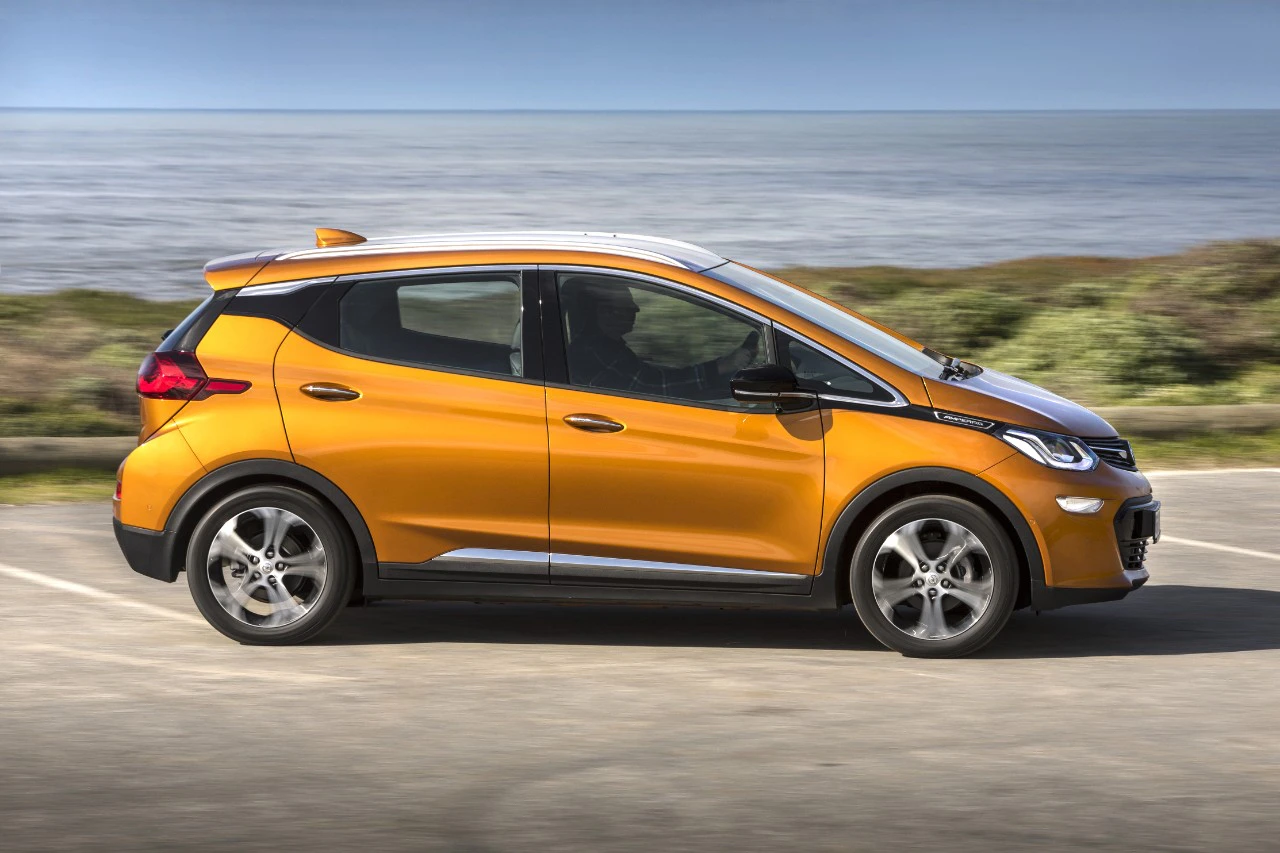 Orangefarbener, kompakter Elektro-SUV fährt auf einer Straße am Meer entlang, umgeben von grünem Gelände und blauem Himmel im Hintergrund.