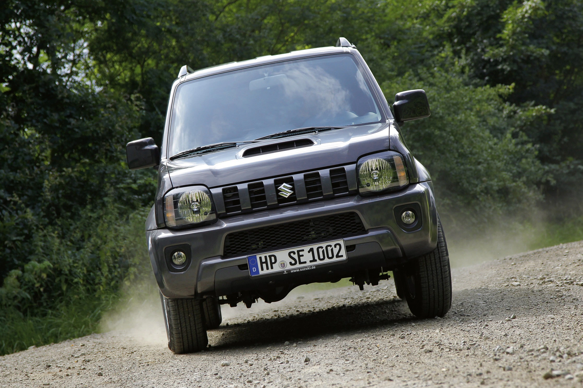 Silberner Suzuki-Geländewagen fährt auf staubigem Weg, umgeben von grünen Bäumen. Frontalansicht mit sichtbarem Nummernschild.