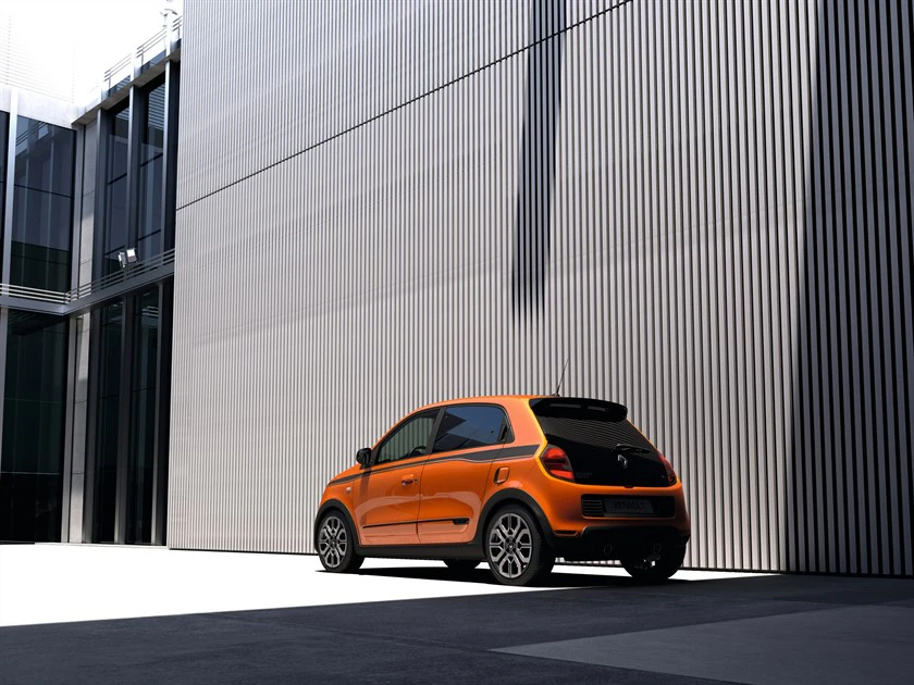 Orangefarbenes Auto parkt vor einem hohen, modernen Gebäude mit Metallfassade und vertikalen Streifen. Sonnenlicht erzeugt starke Schatten.