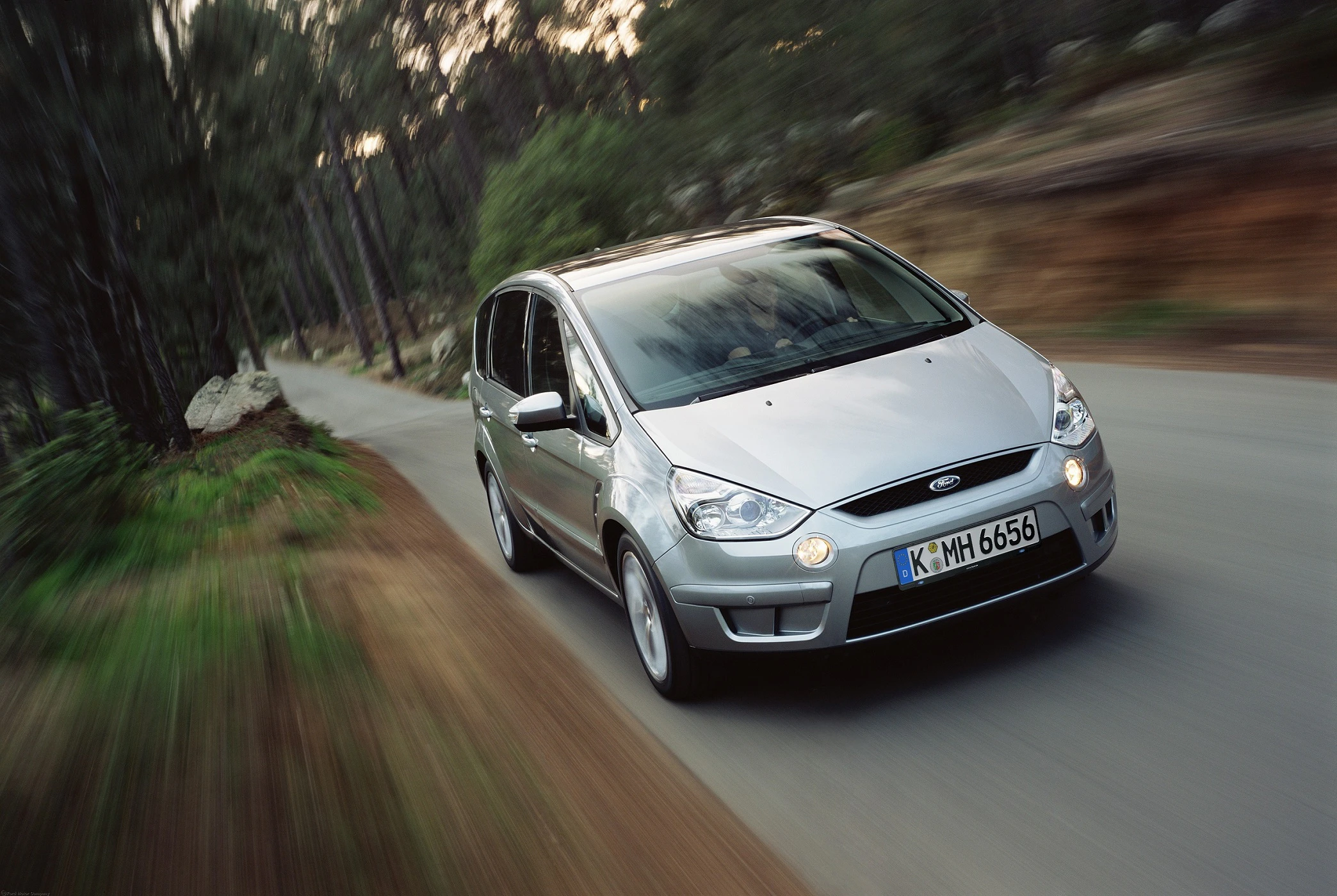 Silberner Ford S-MAX fährt auf einer schmalen, kurvigen Straße durch einen Wald. Geschwindigkeitseffekt betont die Bewegung.