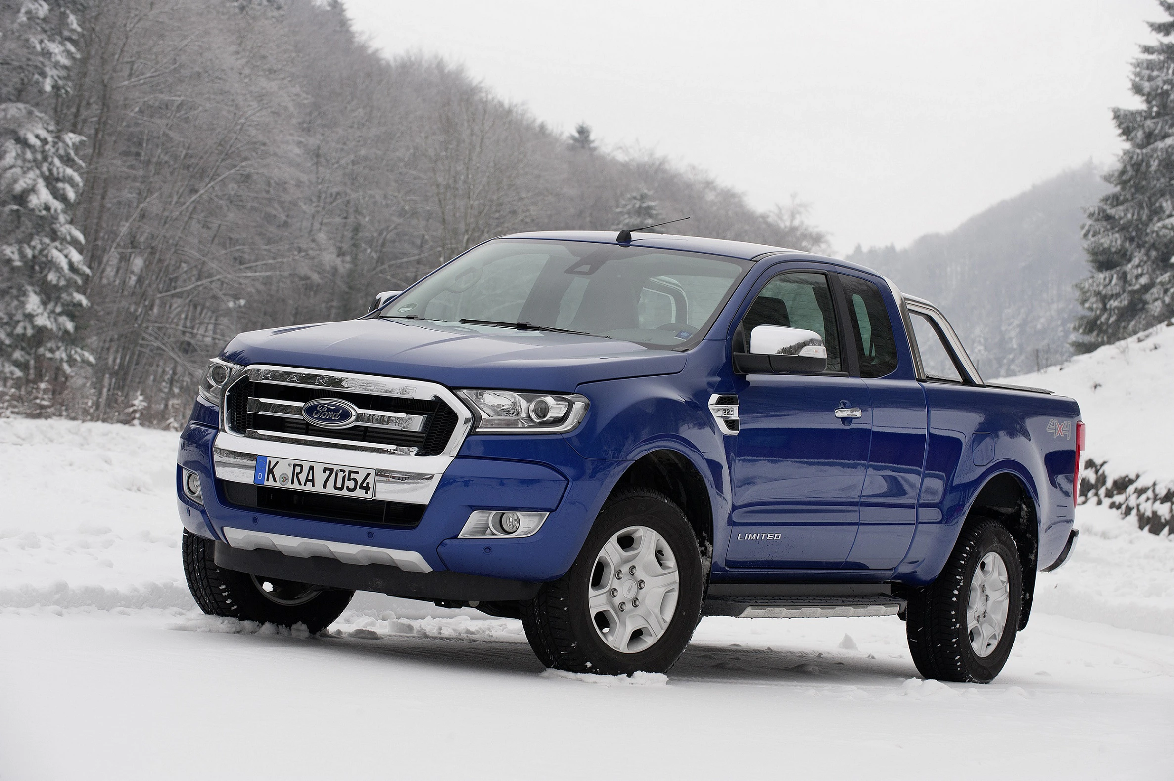 Blauer Pickup-Truck steht auf schneebedecktem Boden vor schneebedecktem Wald. Fahrzeug im Vordergrund, winterliche Landschaft im Hintergrund.