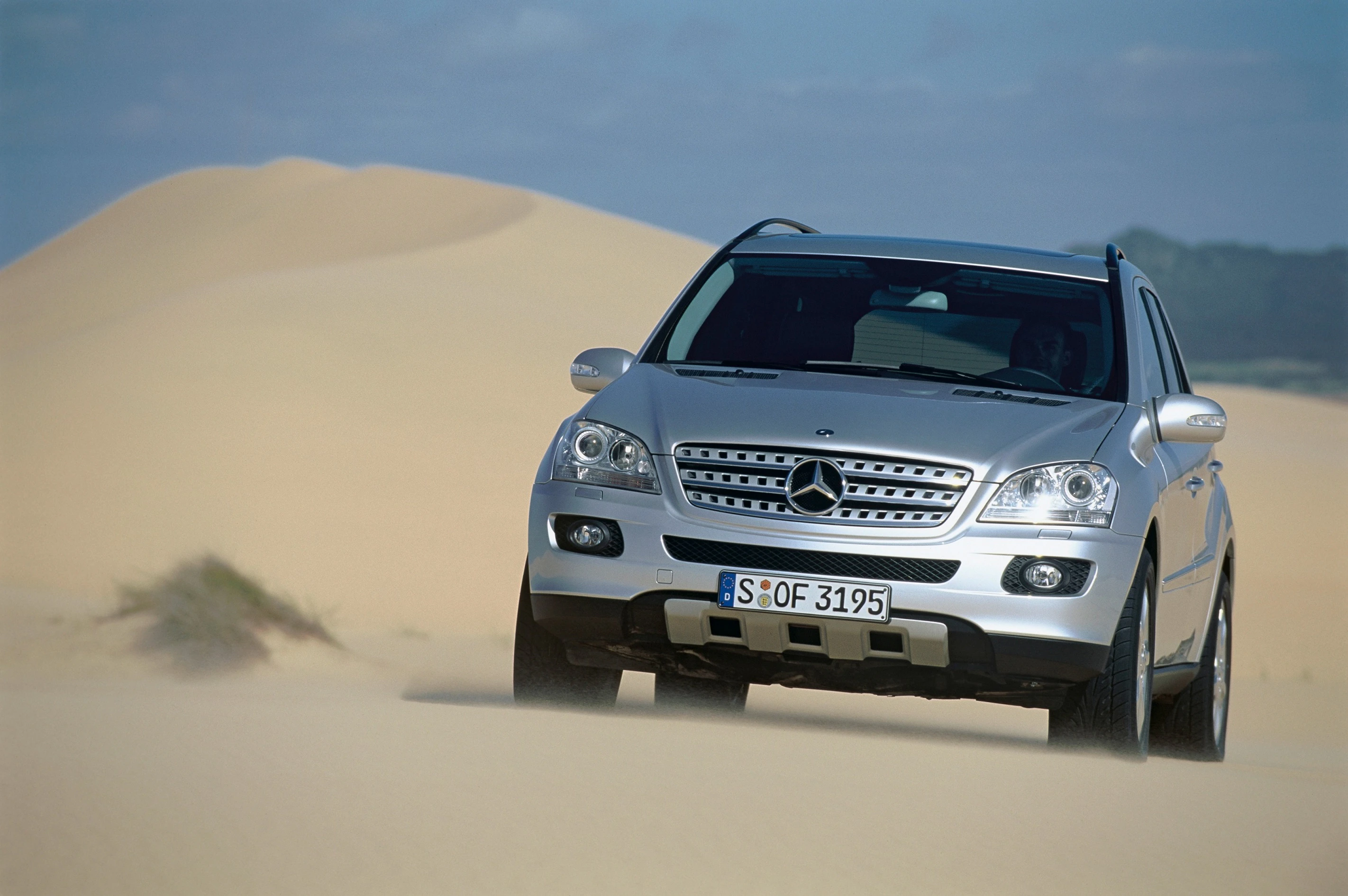 Silberner SUV fährt durch Sanddünen unter blauem Himmel. Staub wirbelt um die Reifen, während das Fahrzeug leicht geneigt ist.