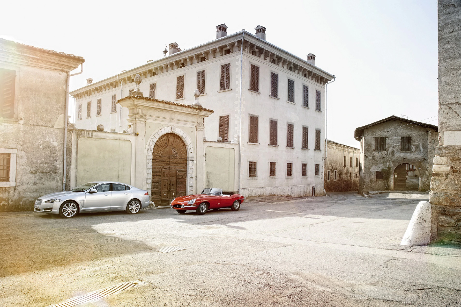 Zwei klassische Autos parken vor einem alten, großen Gebäude mit verziertem Torbogen auf einem sonnigen, verlassenen Platz.