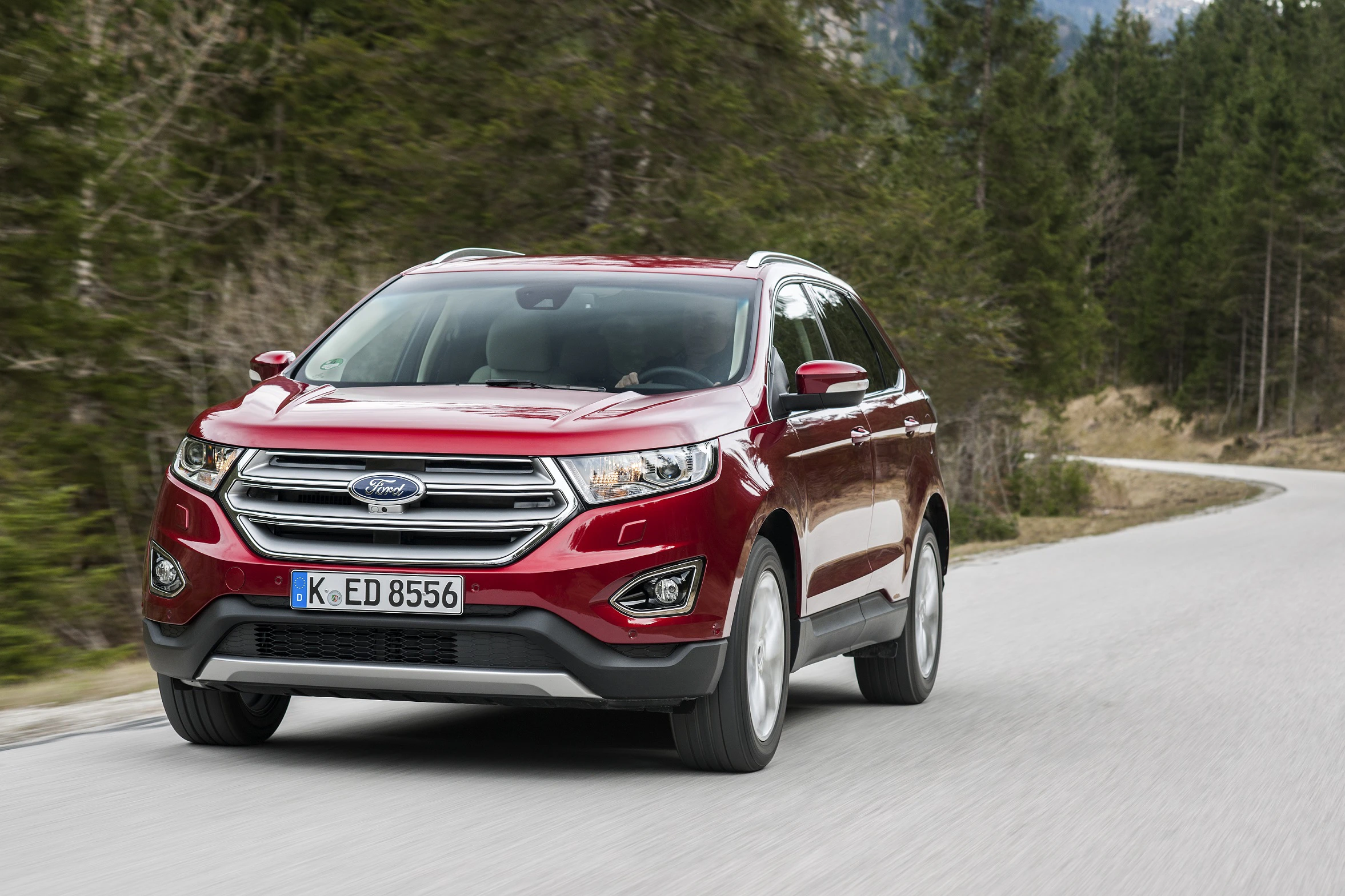 Roter SUV fährt auf einer kurvigen Landstraße, umgeben von Bäumen und Bergen im Hintergrund. Das Fahrzeug stammt von der Marke Ford.