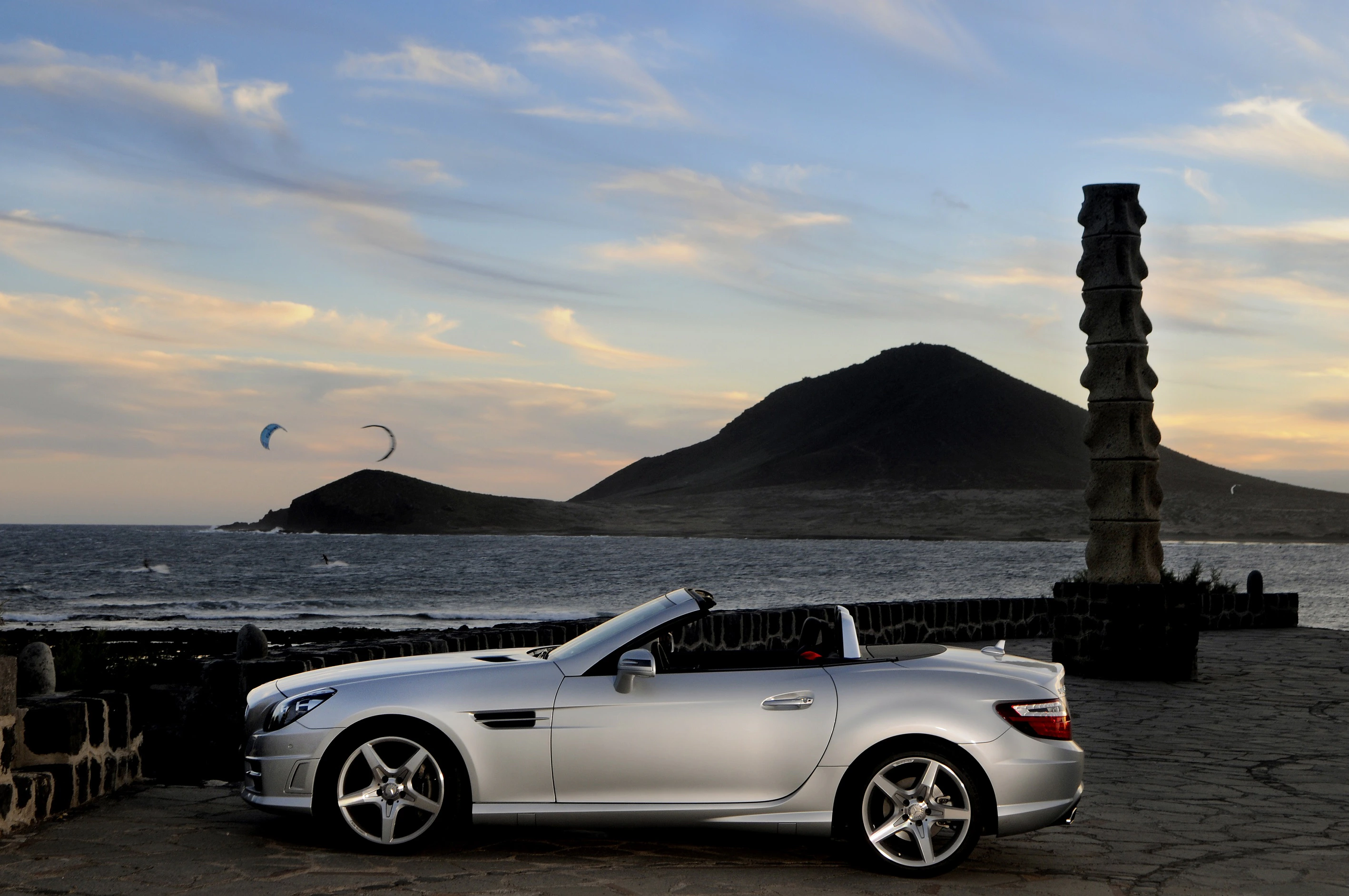 Silbernes Cabriolet vor Küstenlandschaft mit zwei Drachenfliegern am Himmel und einem Hügel im Hintergrund bei Sonnenuntergang.