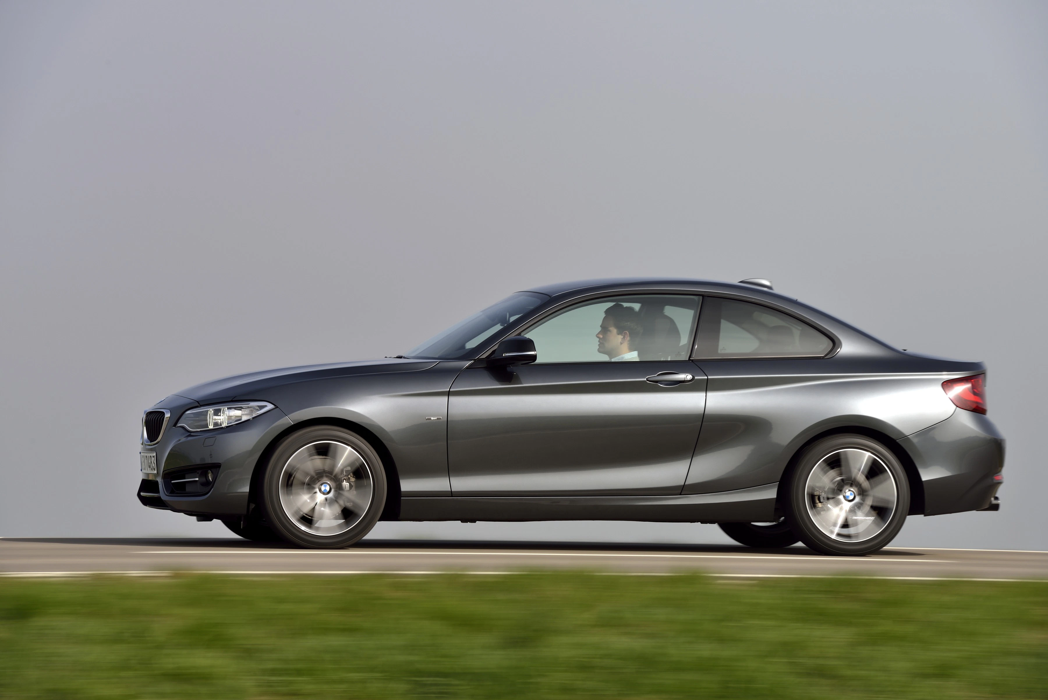 Graues Coupé fährt auf einer Straße vor grauem Himmel; Profilansicht mit sichtbar fahrendem Fahrer.