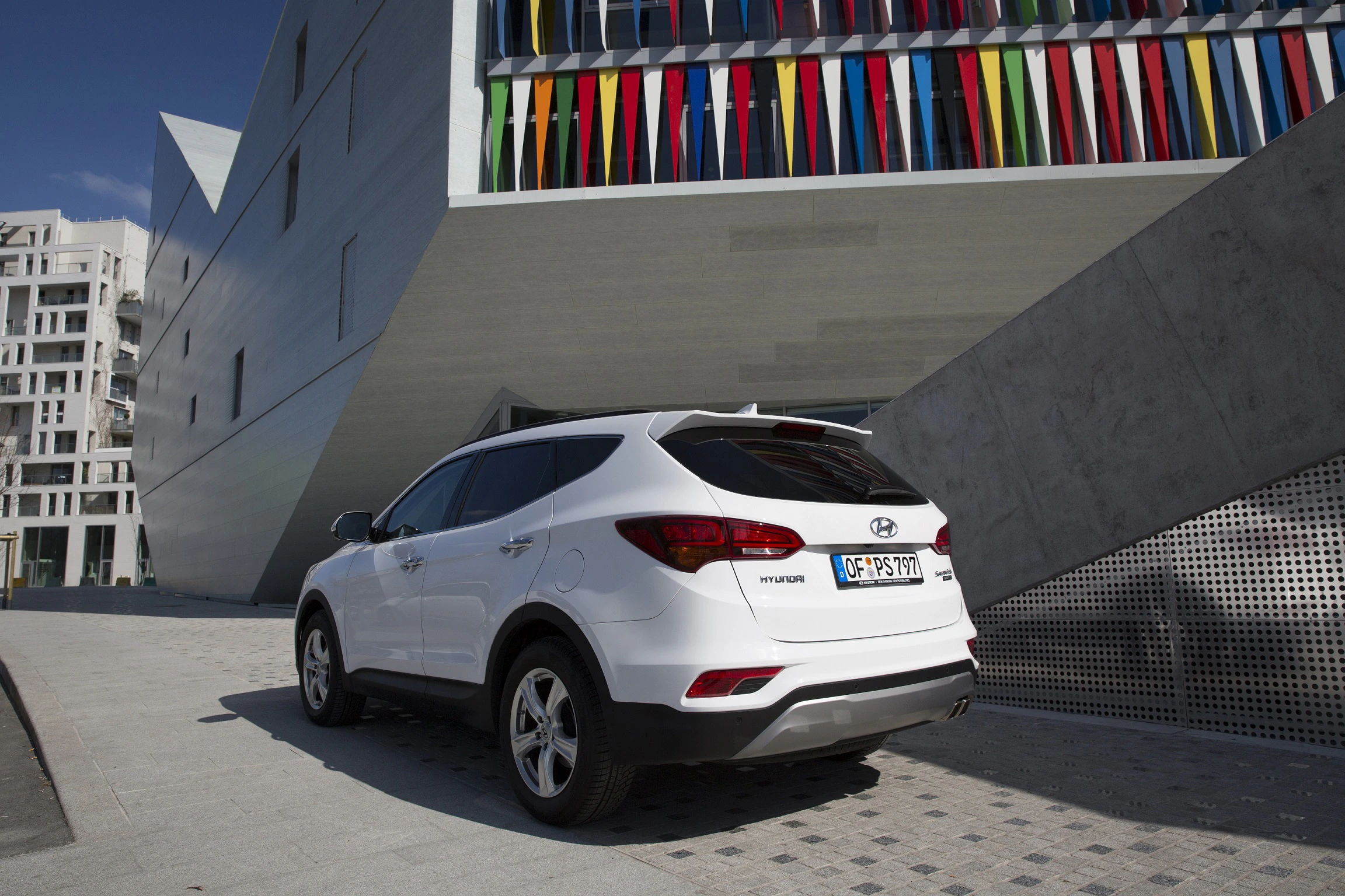Weißer SUV parkt vor einem modernen Gebäude mit bunten Fensterläden und schräger Architektur.