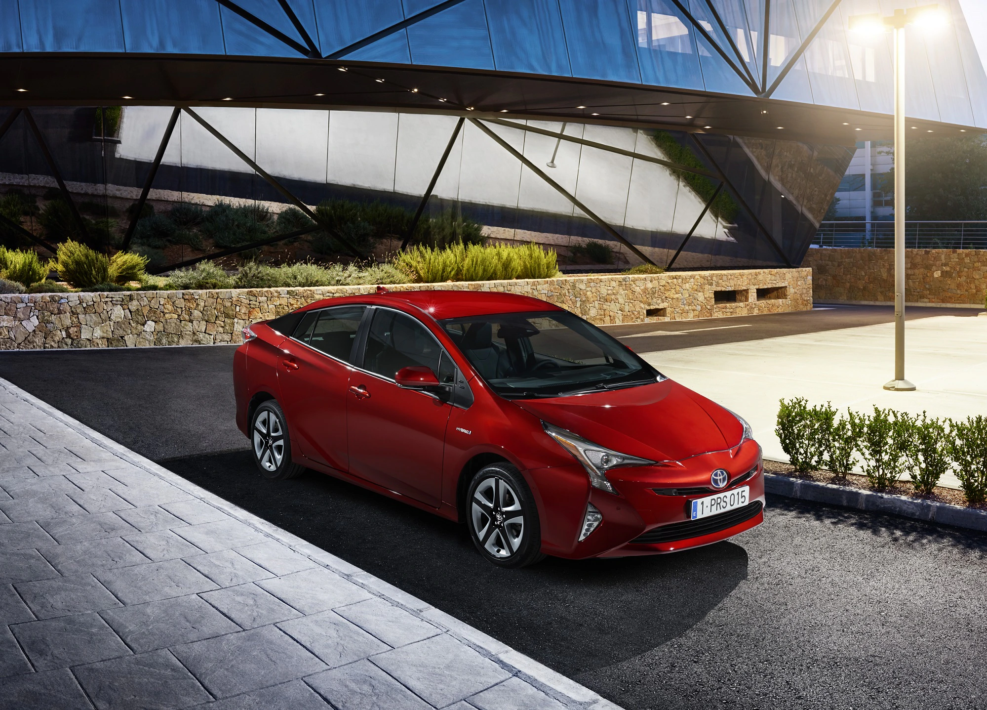 Roter Toyota Prius parkt vor einem modernen Gebäude mit Glasfassade und Pflanzen, abends beleuchtet bei Straßenlaternenlicht.