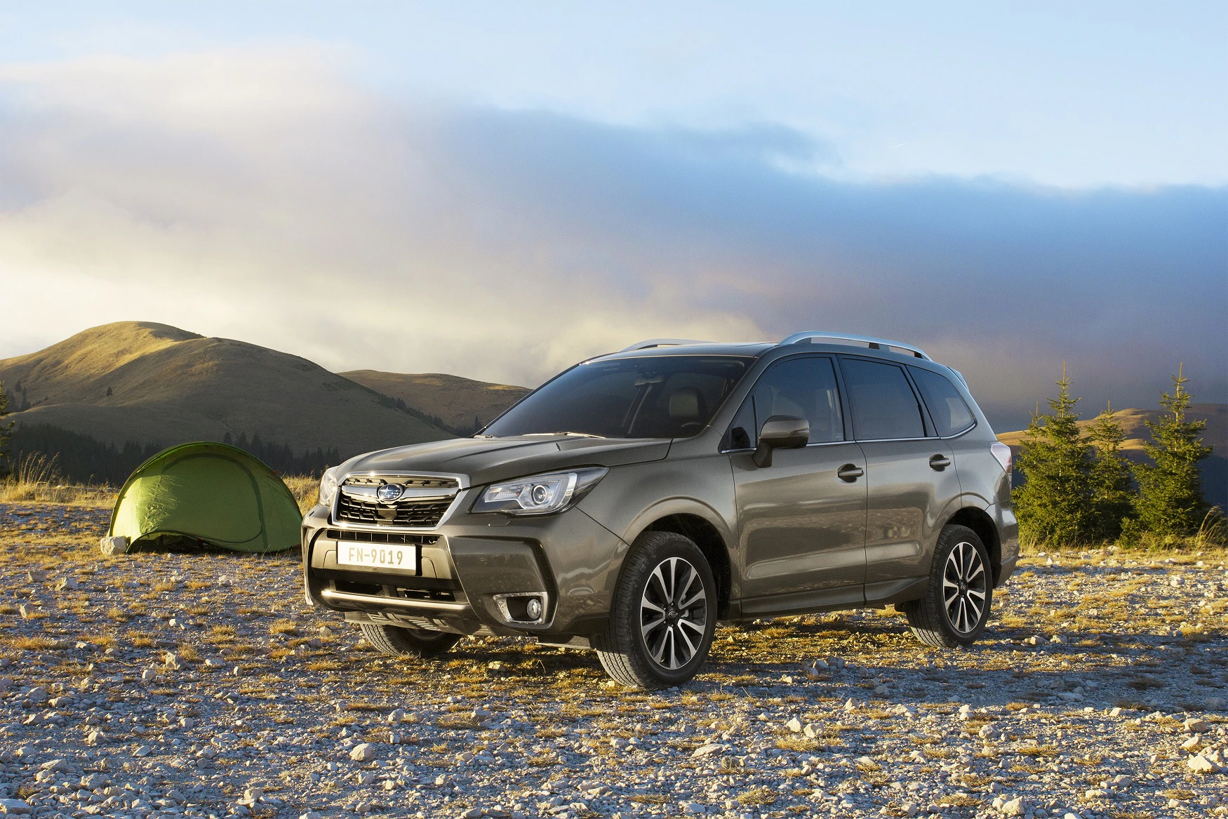 Ein grauer SUV steht auf einem Kiesplatz in einer bergigen Landschaft. Im Hintergrund ist ein Zelt und bewaldete Hügel zu sehen.
