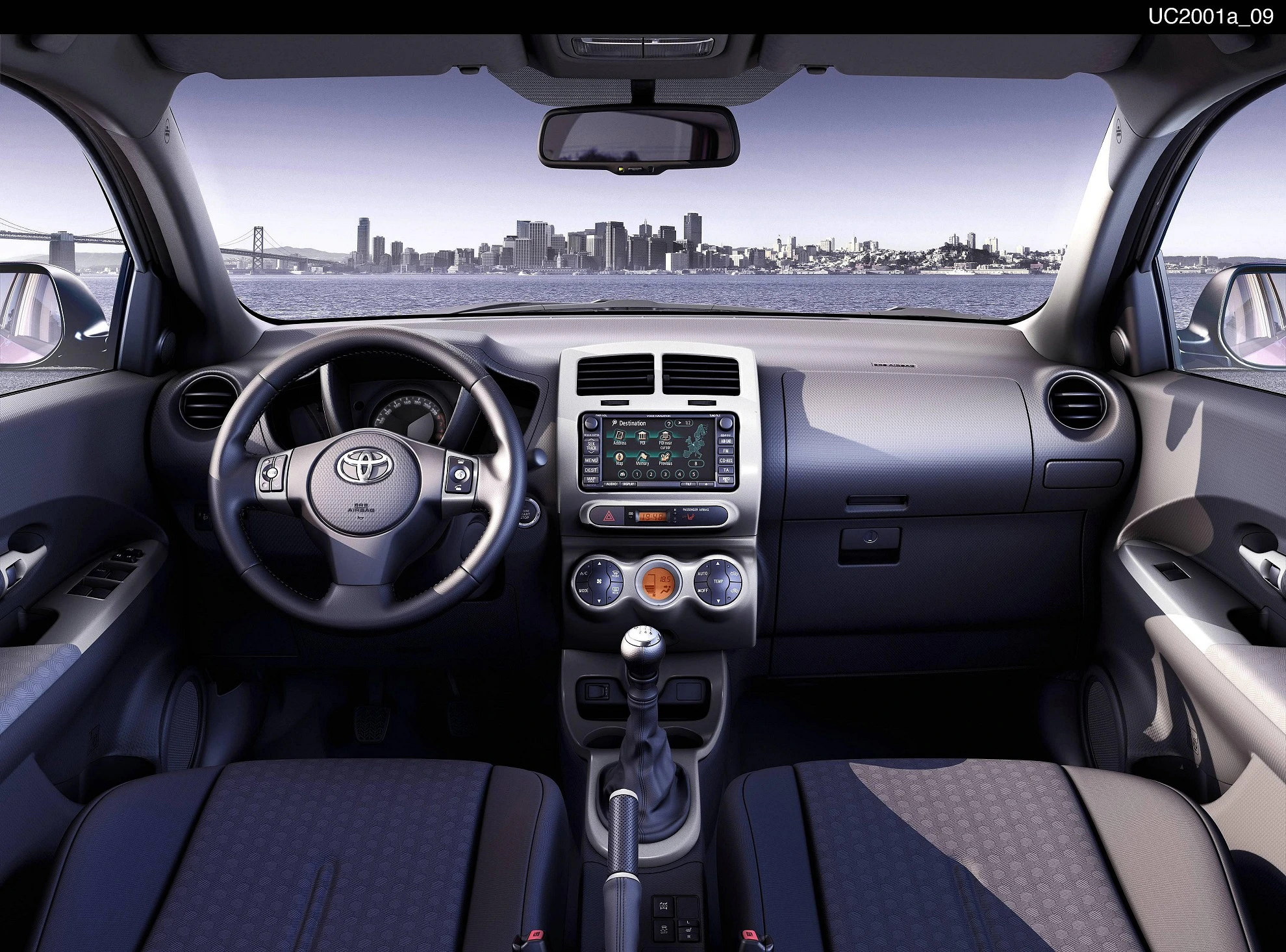 Innenraumansicht eines Autos mit Blick durch Frontscheibe auf eine Stadtlandschaft. Armaturenbrett mit Lenkrad, Schalthebel und Touchscreen.