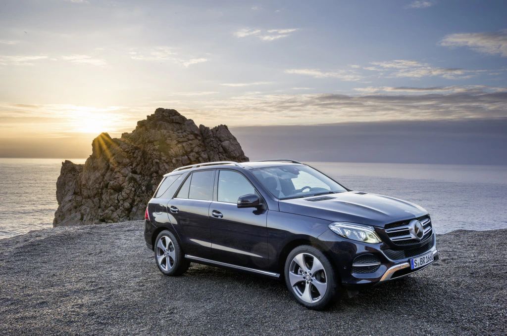 Mercedes-SUV parkt auf einer felsigen Küstenklippe bei Sonnenuntergang. Im Hintergrund das Meer und ein großer Felsen.