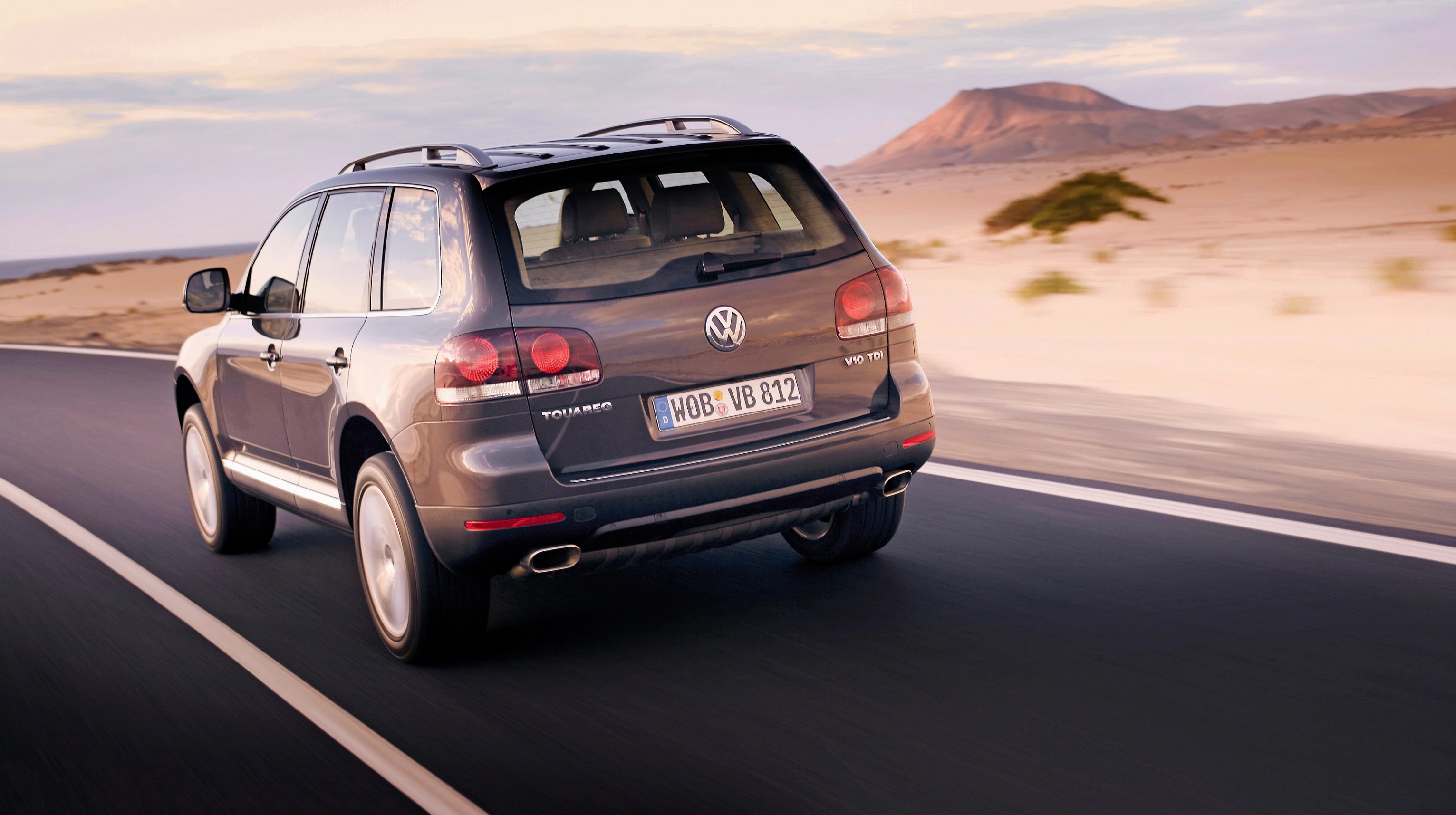 SUV fährt auf einer Straße durch eine Wüstenlandschaft, Berge im Hintergrund, unter einem bewölkten Himmel.