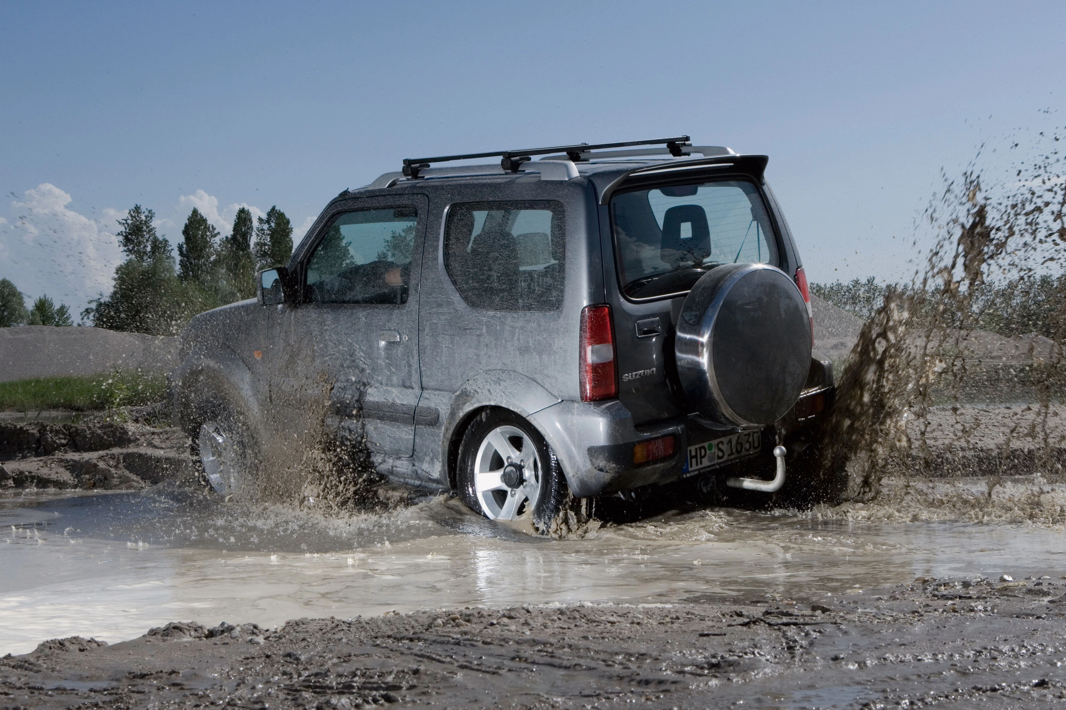 Ein silberner Geländewagen fährt durch eine schlammige Wasserpfütze auf einer unbefestigten Straße bei klarem Himmel.