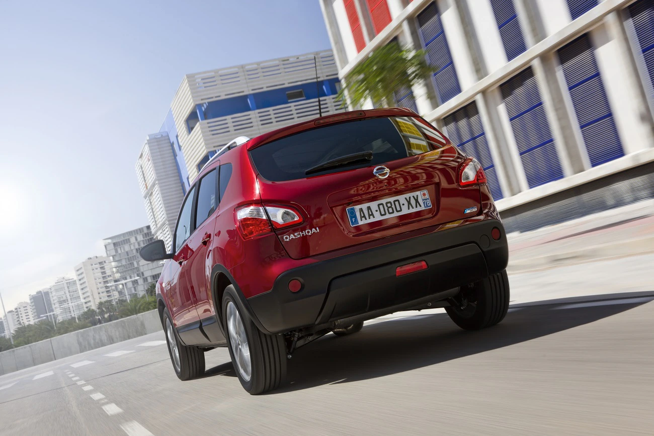 Roter Dacia Qashqai fährt in einer modernen Stadt mit hohen Gebäuden. Man sieht das Auto von hinten auf der Straße.