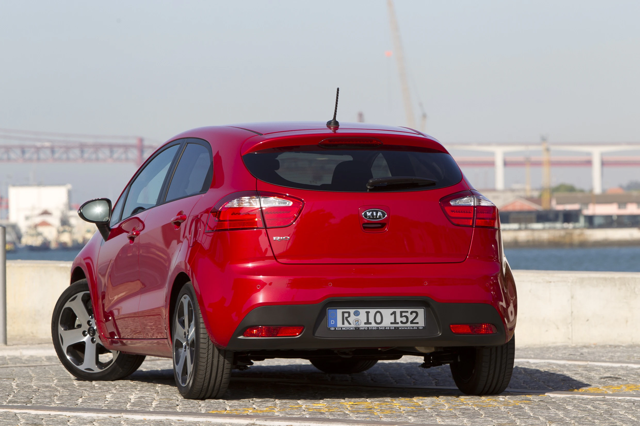 Roter Kia Rio parkt auf Kopfsteinpflaster vor einem Fluss, im Hintergrund ist eine große Brücke sichtbar.