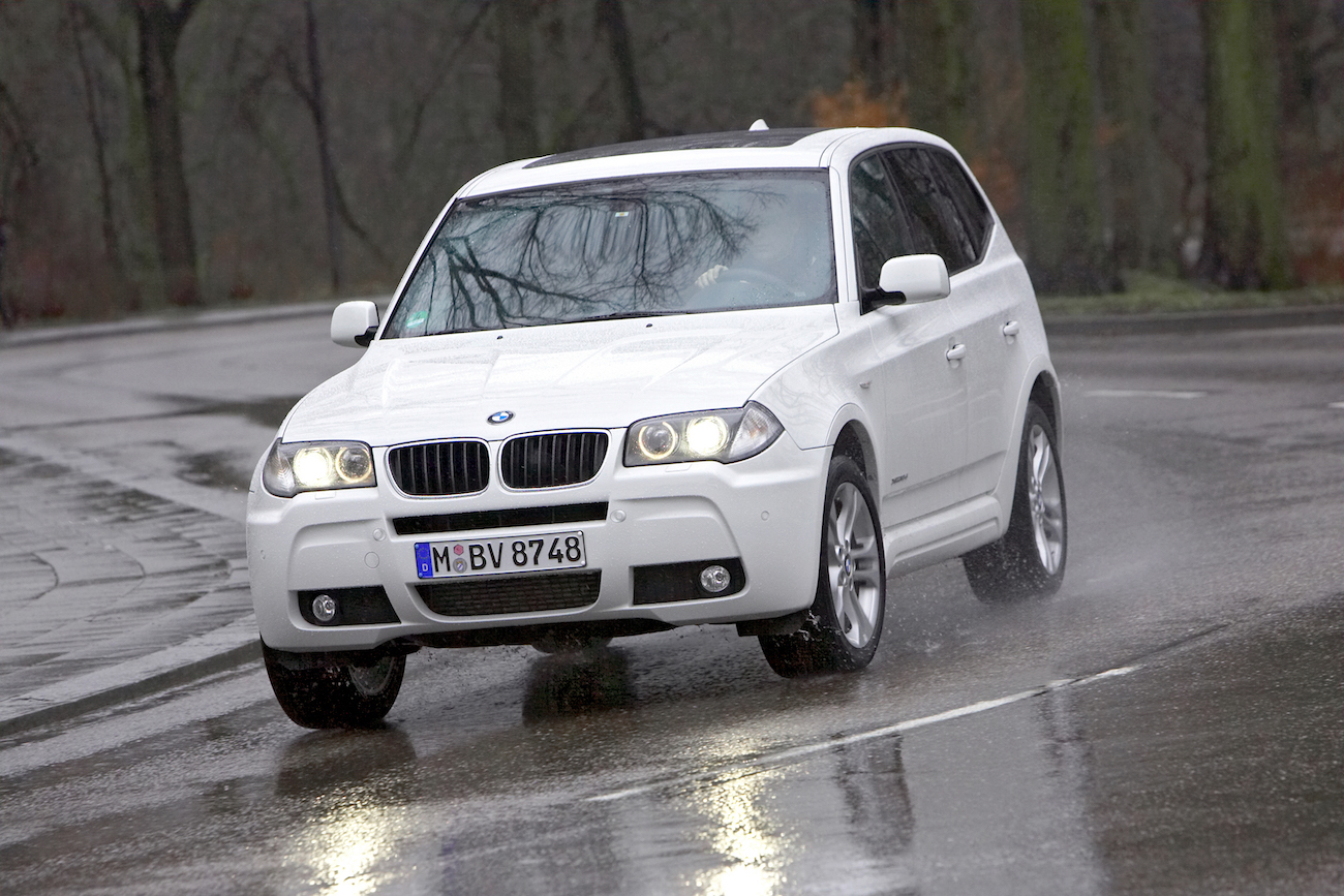 Ein weißer BMW X3 fährt bei Regen auf einer nassen Straße, Wasser spritzt von den Reifen.