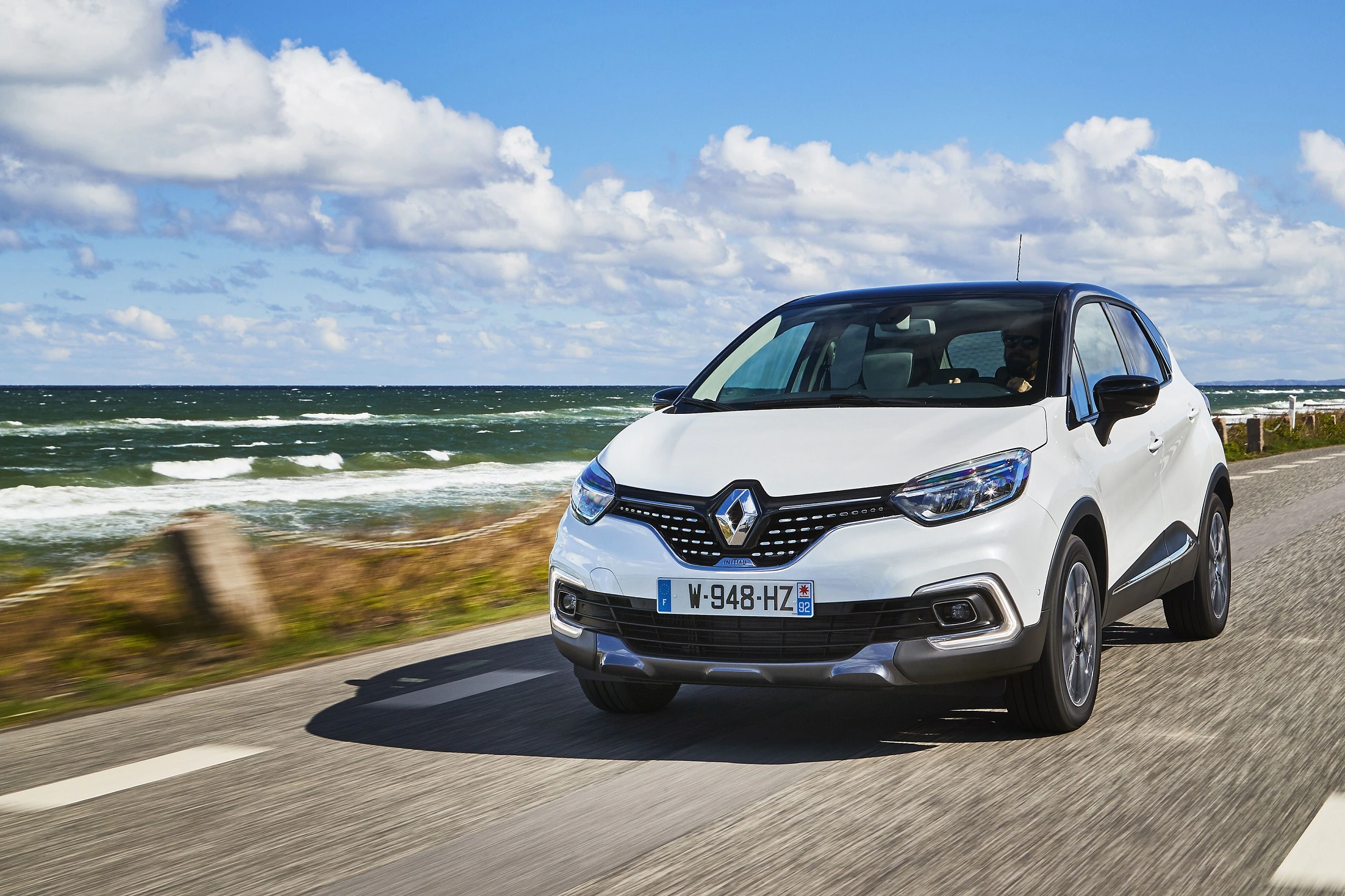Weißer Renault fährt auf einer Küstenstraße bei sonnigem Wetter. Im Hintergrund das Meer und ein blauer Himmel mit Wolken.