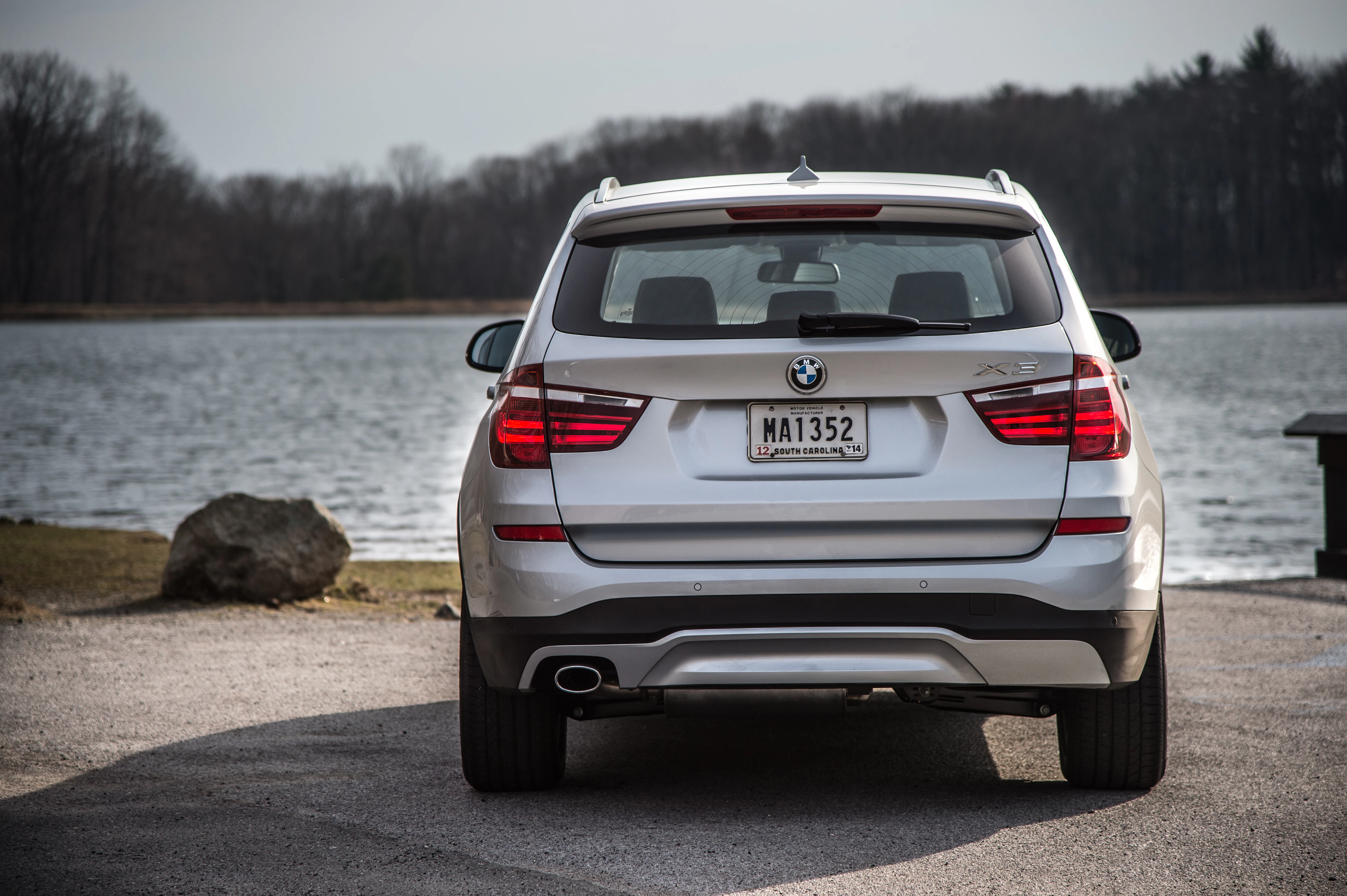 Silberner BMW X3 SUV steht am Ufer eines Sees, umgeben von kahlen Bäumen, auf einem asphaltierten Parkplatz bei Tageslicht.