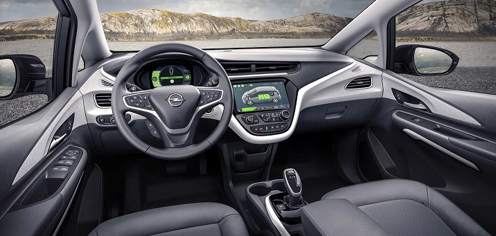 Innenraum eines modernen Autos mit grauen Sitzen, digitalem Armaturenbrett und Lenkrad, umgeben von Felsenlandschaft im Hintergrund.