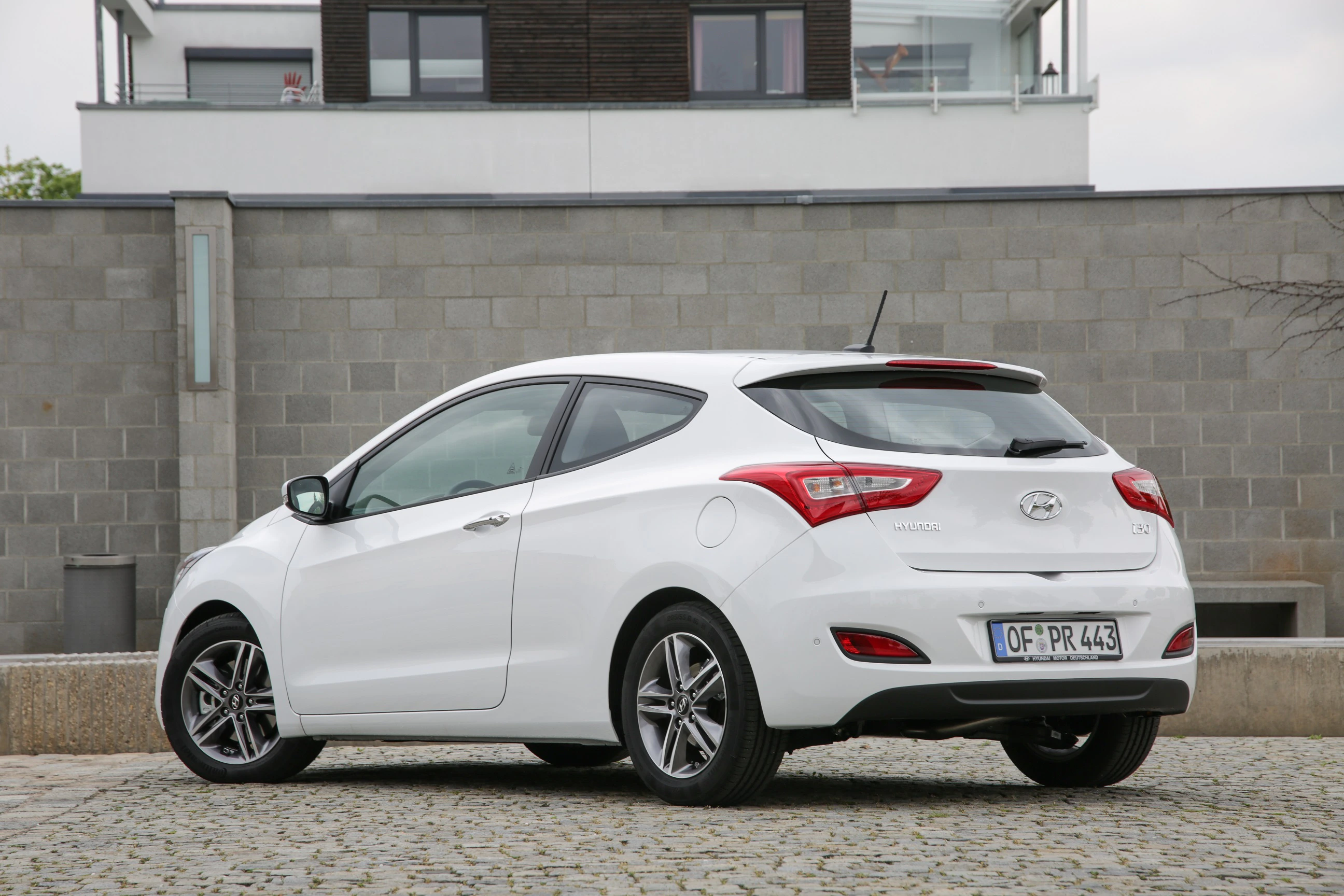 Weißer Hyundai i30 vor einer grauen Mauer, auf einem gepflasterten Platz geparkt. Ansicht von hinten links.