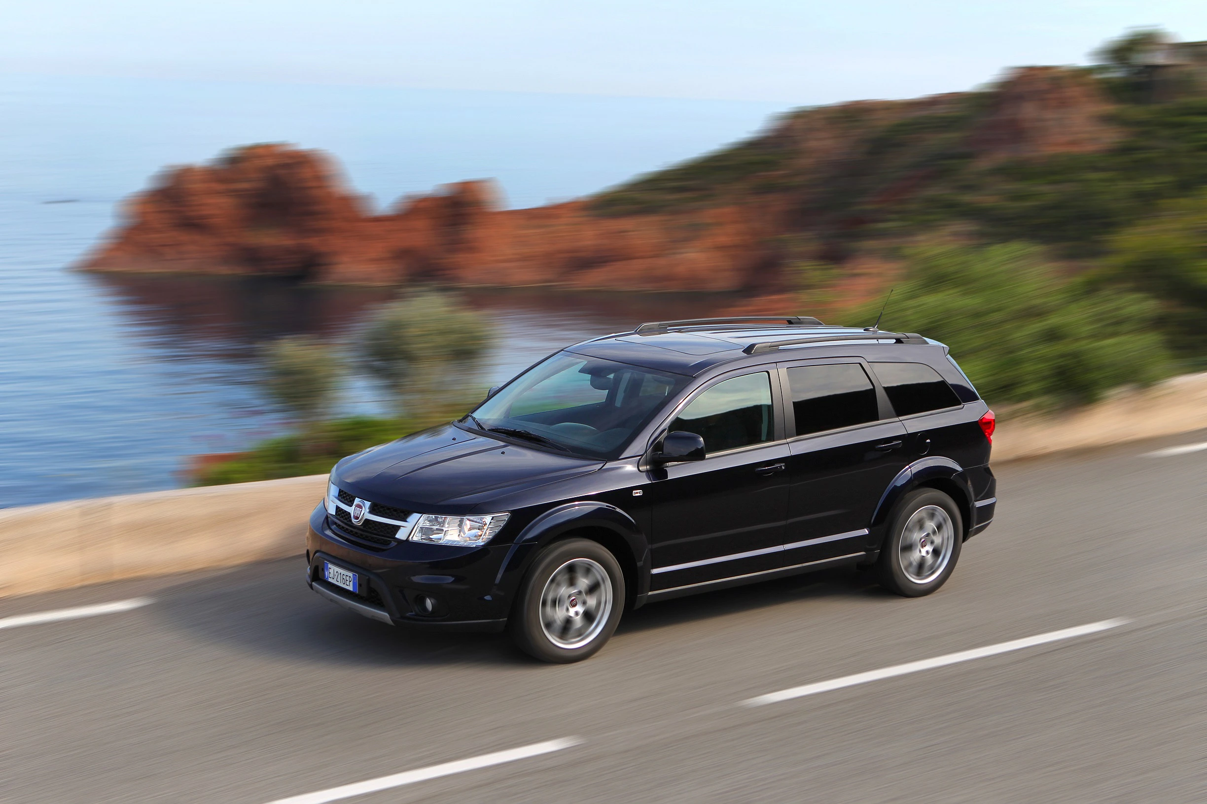 Schwarzer SUV fährt auf Küstenstraße mit roter Felslandschaft und blauem Meer im Hintergrund.