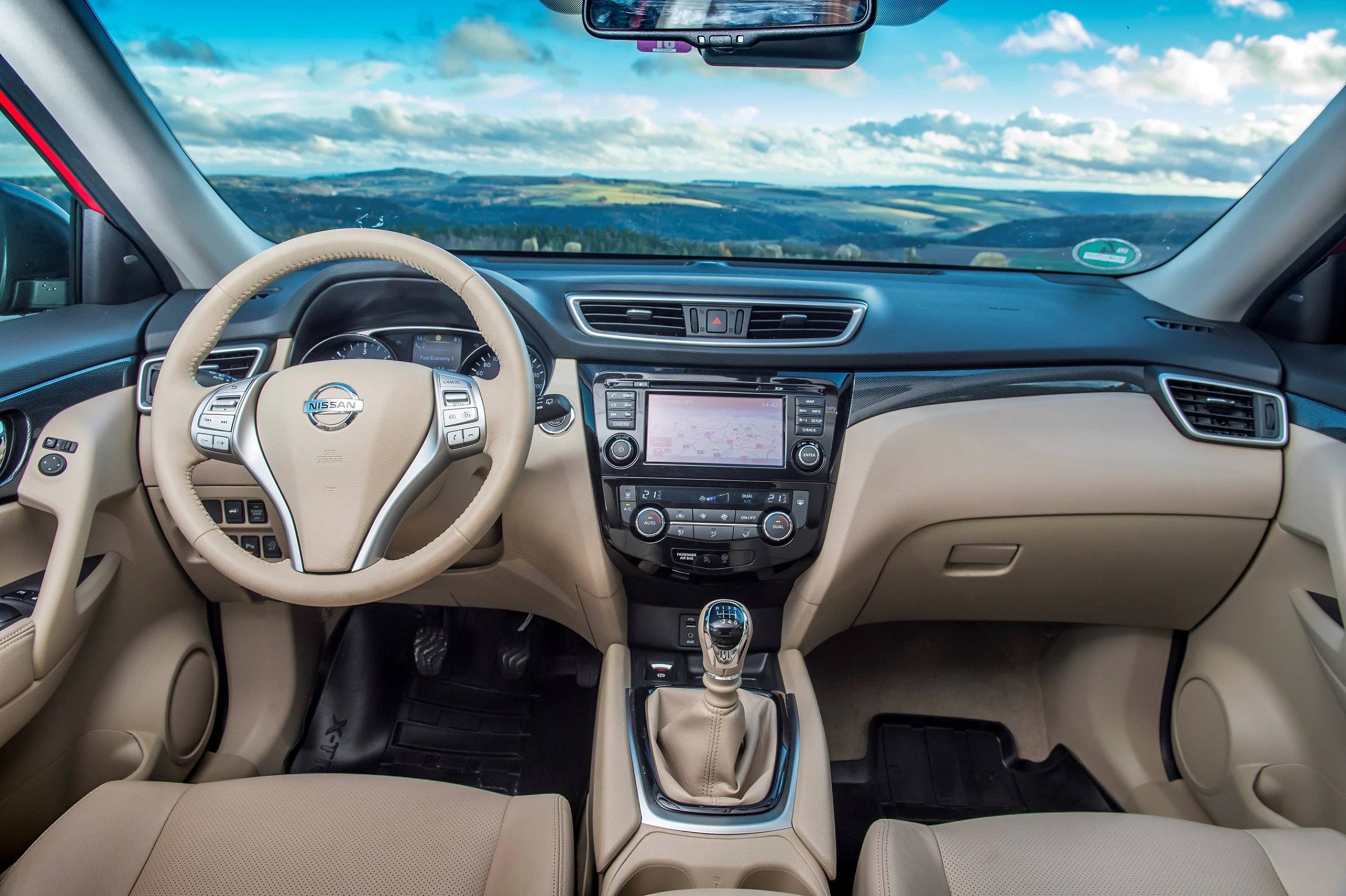 Innenraum eines beige-schwarzen Autos mit Lenkrad, Schalthebel und Infotainment-System. Landschaft durch die Windschutzscheibe sichtbar.