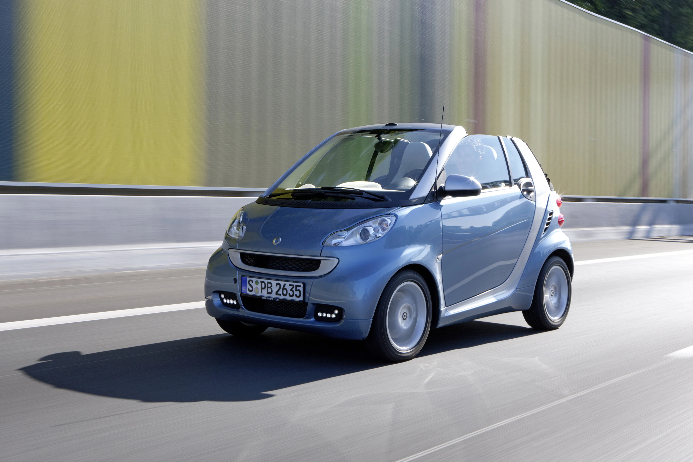 Blauer Smart Fortwo Cabrio fährt auf einer Straße entlang einer farbigen Lärmschutzwand.