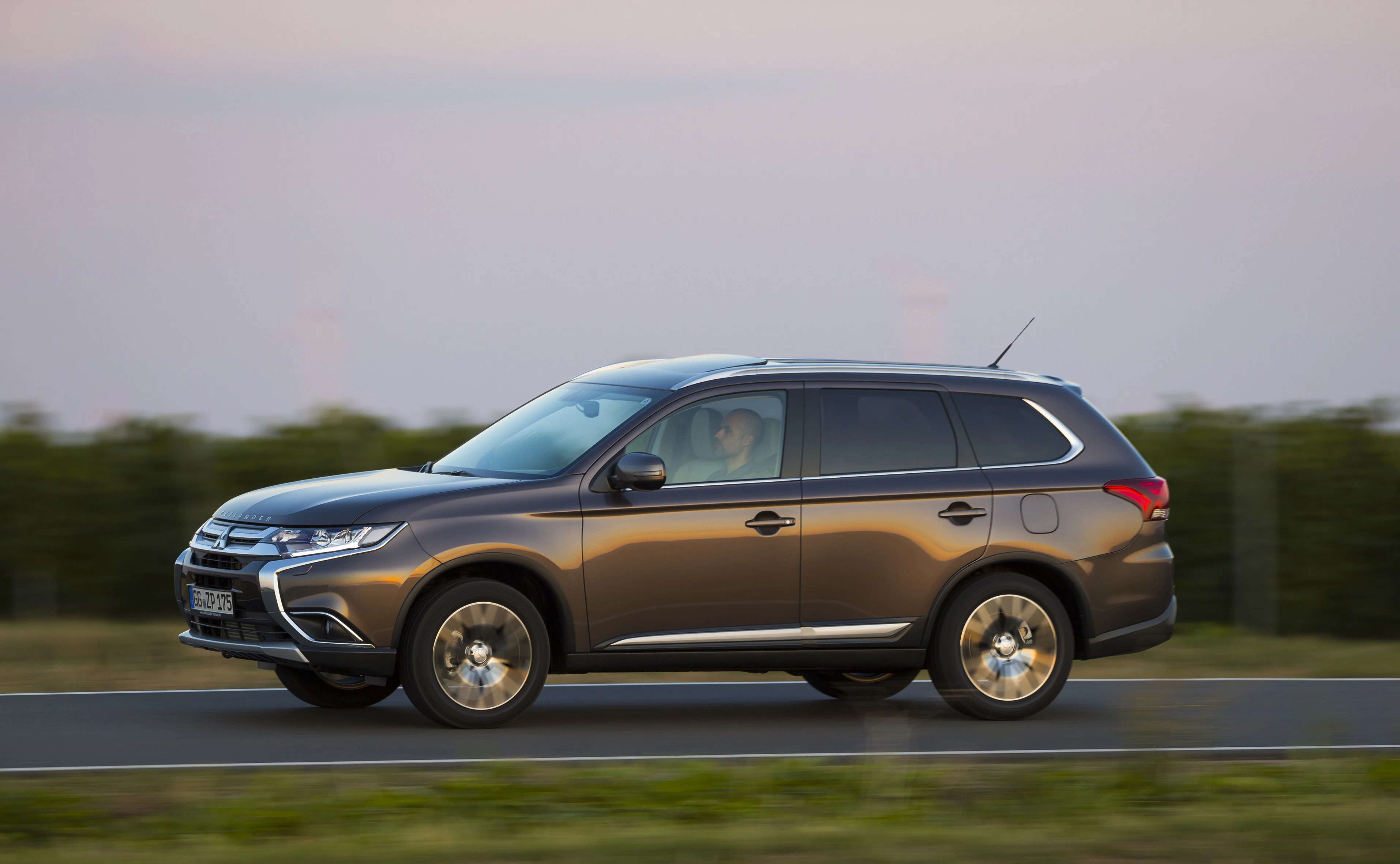 Autofahrt in der Abenddämmerung: Ein grauer SUV fährt auf einer Landstraße, umgeben von grüner Vegetation. Der Himmel ist rosa gefärbt.