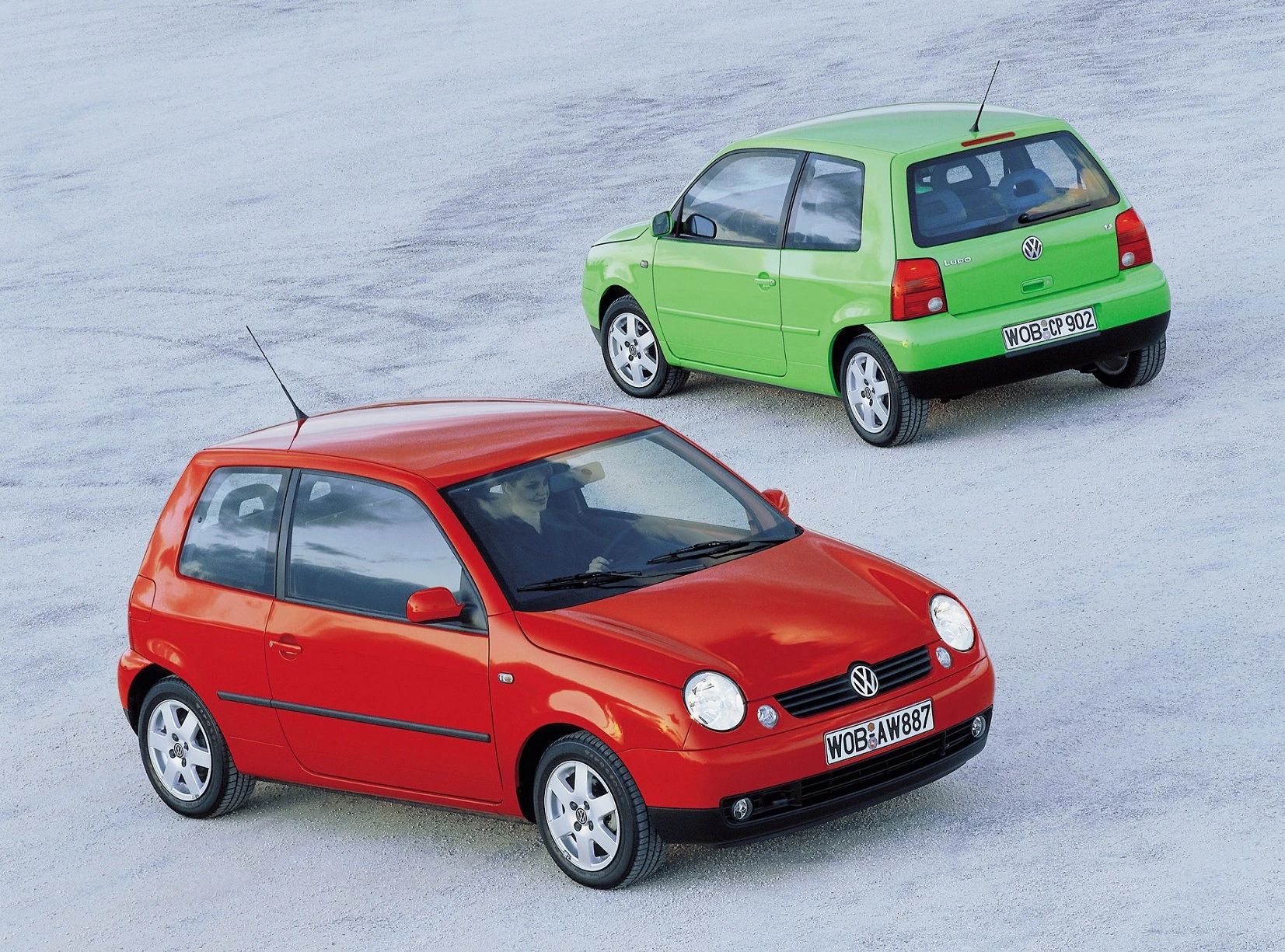 Zwei farbige Autos von Volkswagen auf einer hellen Oberfläche geparkt; ein roter und ein grüner Wagen in einem leichten Winkel zueinander.